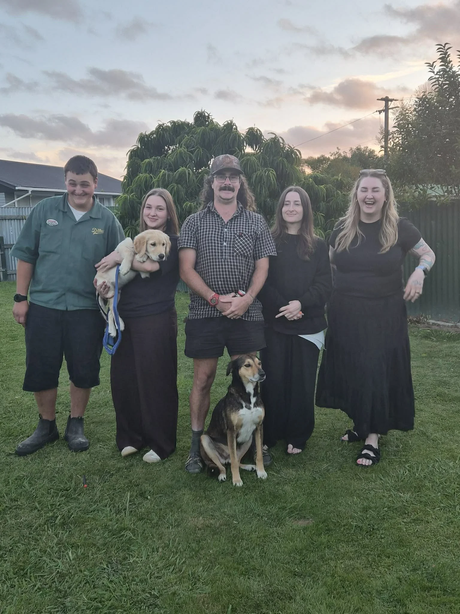 A group of five people and two dogs standing outdoors on grass during sunset, with trees, a house, and power lines in the background.