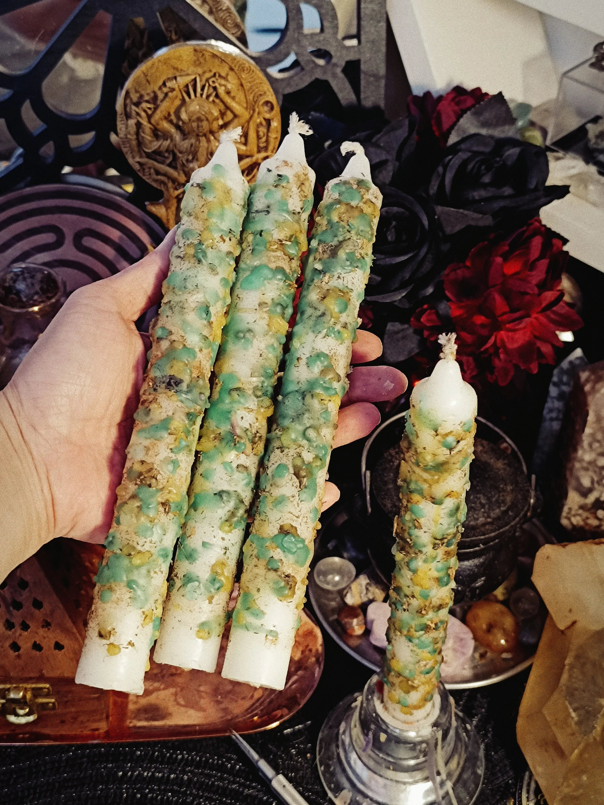 Handmade white prosperity taper candles with green and gold wax drips and herbs, shown in hand