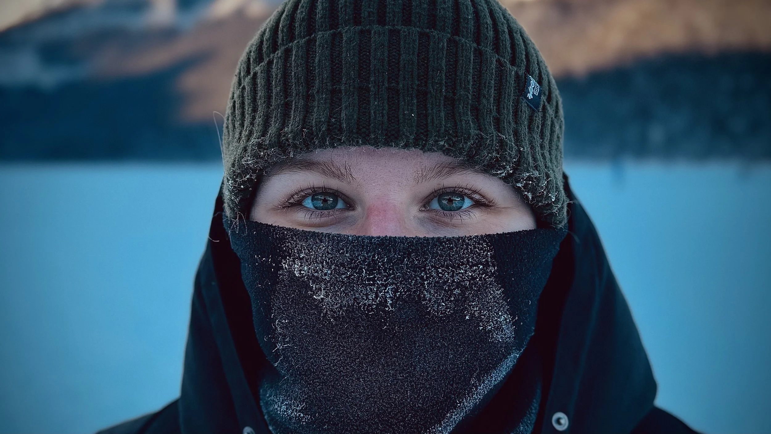 Ragazzo con cappello di lana e passamontagna, con neve sulla faccia e sugli indumenti, in ambiente invernale.