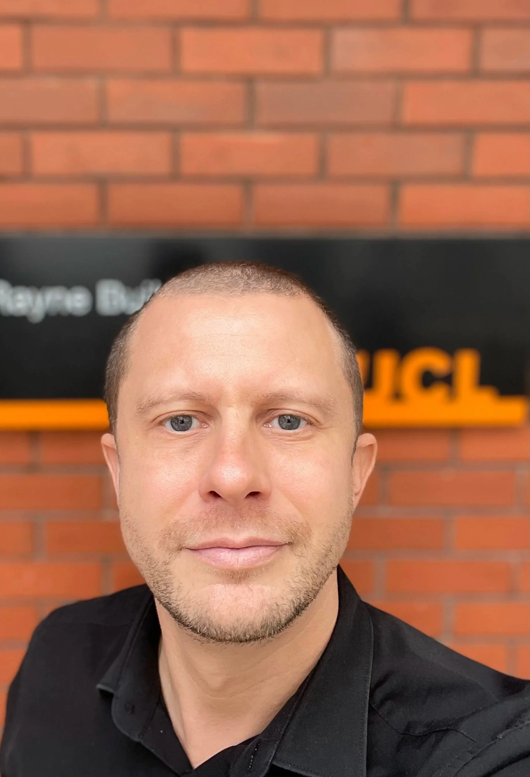 A man taking a selfie in front of a brick wall with a black sign behind him that has white and orange text.