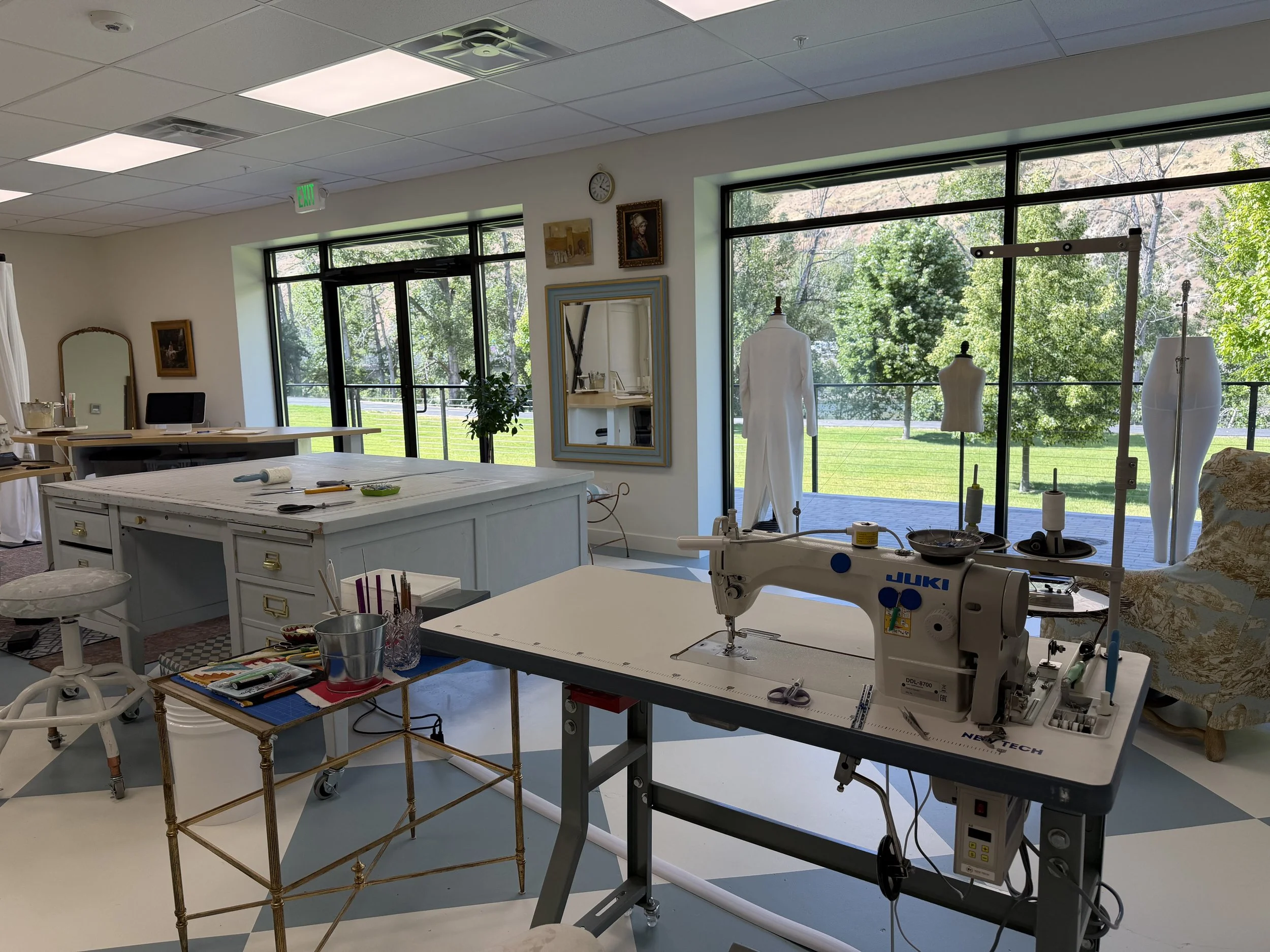 Interior view of a bright room with large windows showing green trees outside; the room contains sewing equipment, mannequins, a mirror, a desk with a computer, and various art supplies.