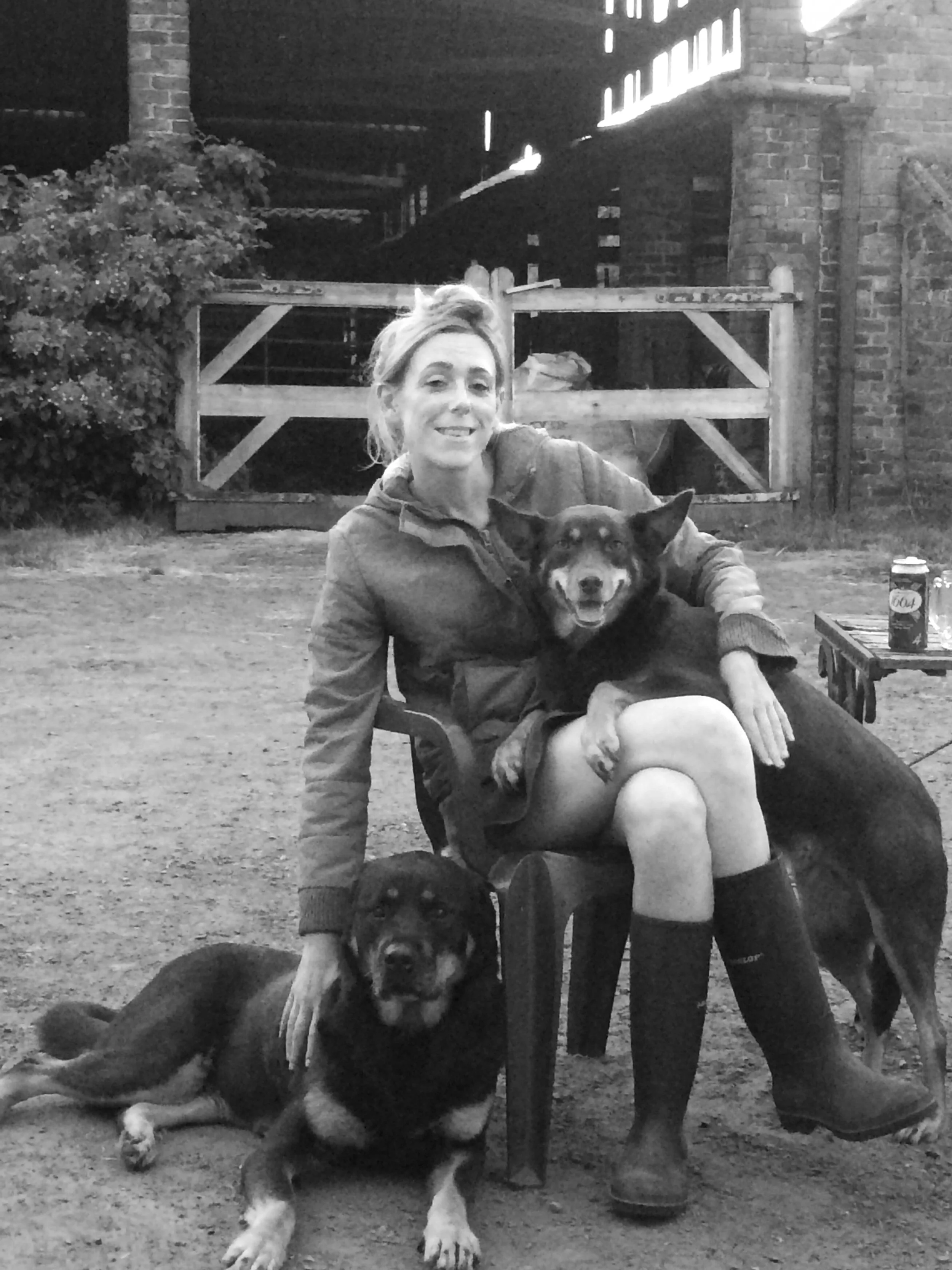 A woman sitting on a chair outdoors with two dogs, one sitting beside her and one lying on the ground. The woman is smiling, wearing a jacket and boots in a rustic setting with wooden structures and brick walls in the background.
