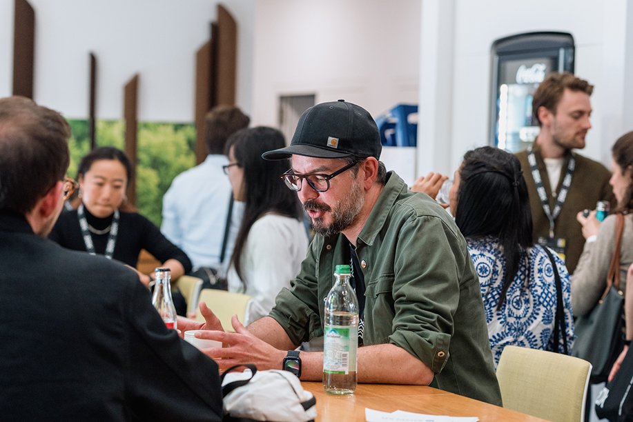 A man with glasses, a black cap, and a green jacket sitting at a table, engaged in conversation, with several people standing and talking in the background at a busy indoor event.
