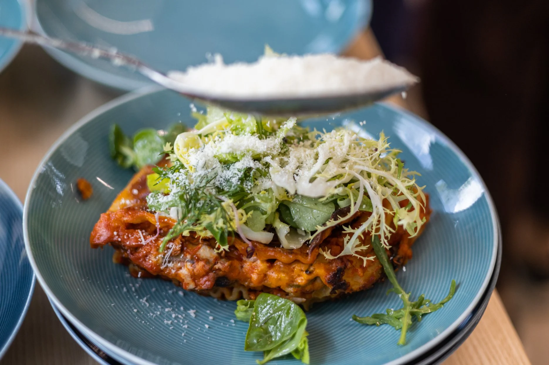 A close-up of lasagna topped with mixed greens and grated cheese on a blue plate.