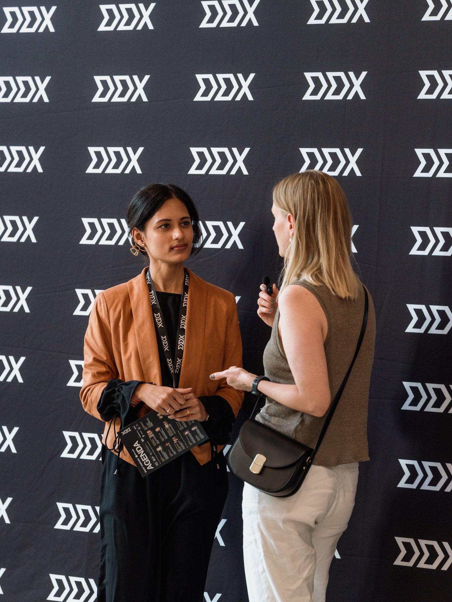 Two women engaged in conversation at a conference, standing in front of a black backdrop with white logos. One woman has dark hair, wears a brown blazer, and holds a brochure. The other has blonde hair, wears a sleeveless top, and holds a microphone.