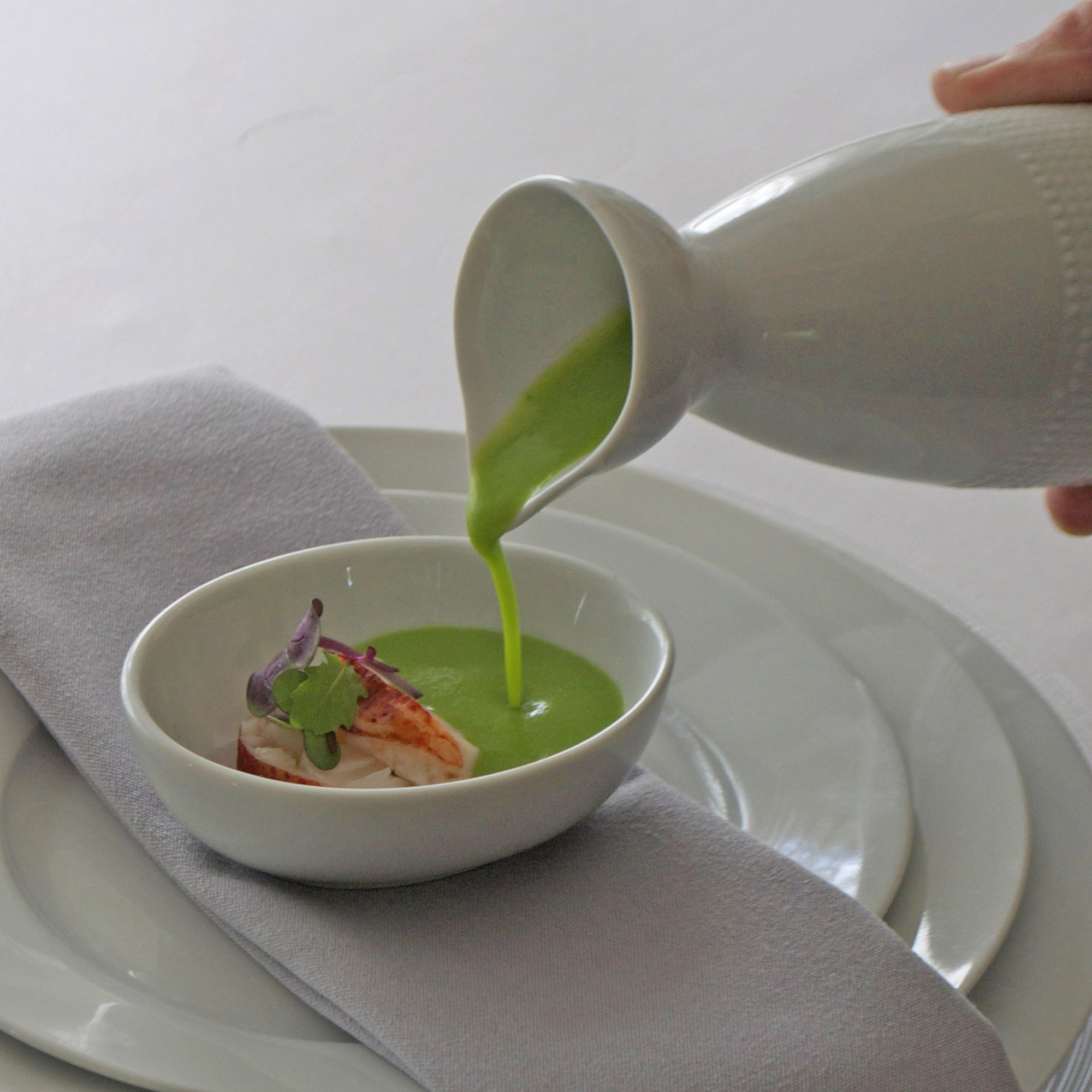 A white cup with a green liquid being poured into it, placed on a white surface with a light grey cloth underneath.