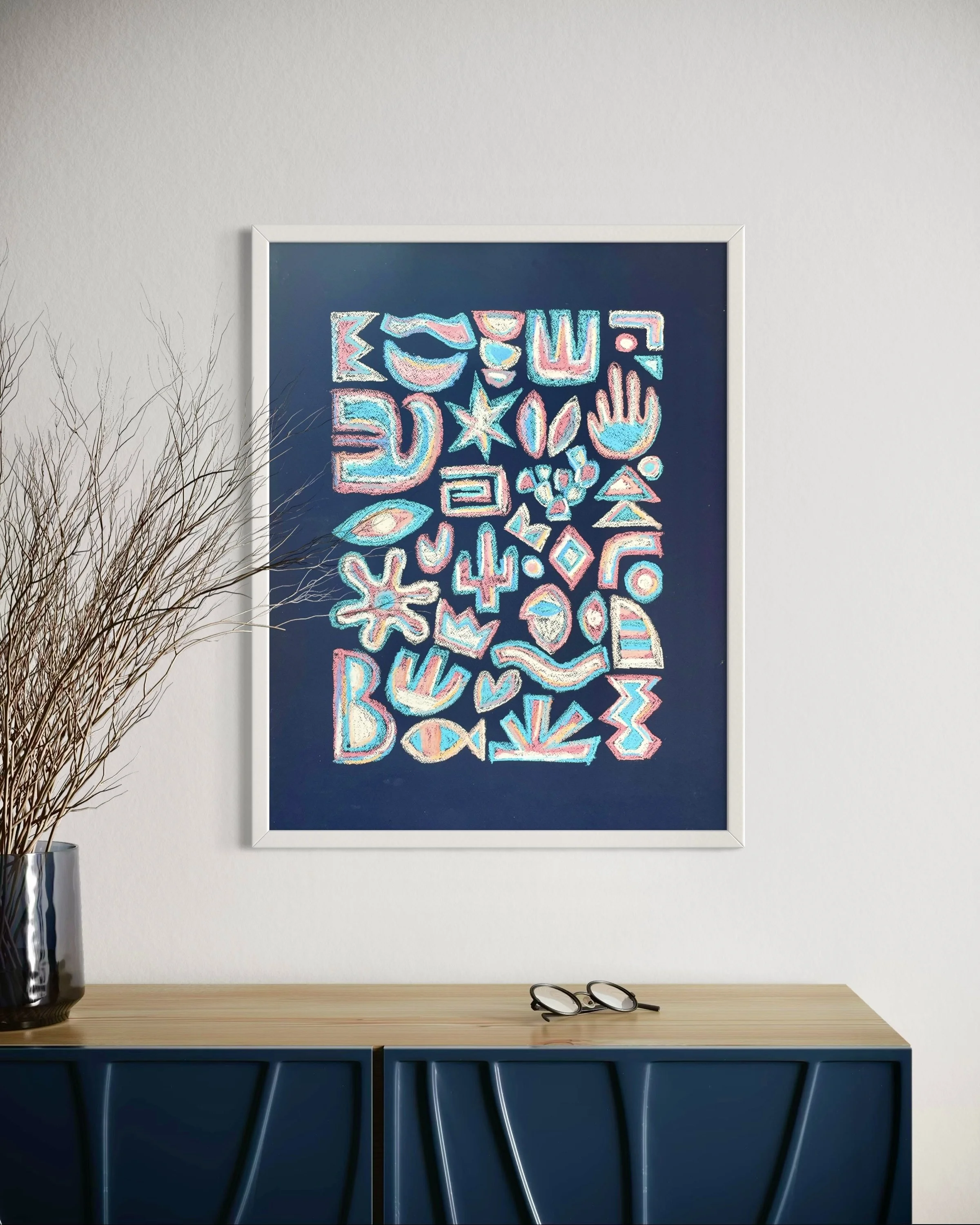 A framed colorful artwork with abstract shapes hangs on a white wall above a wooden desk. On the desk, there is a pair of black-rimmed eyeglasses and a tall silver vase with brown decorative branches. The bottom of the desk is dark blue with vertical
