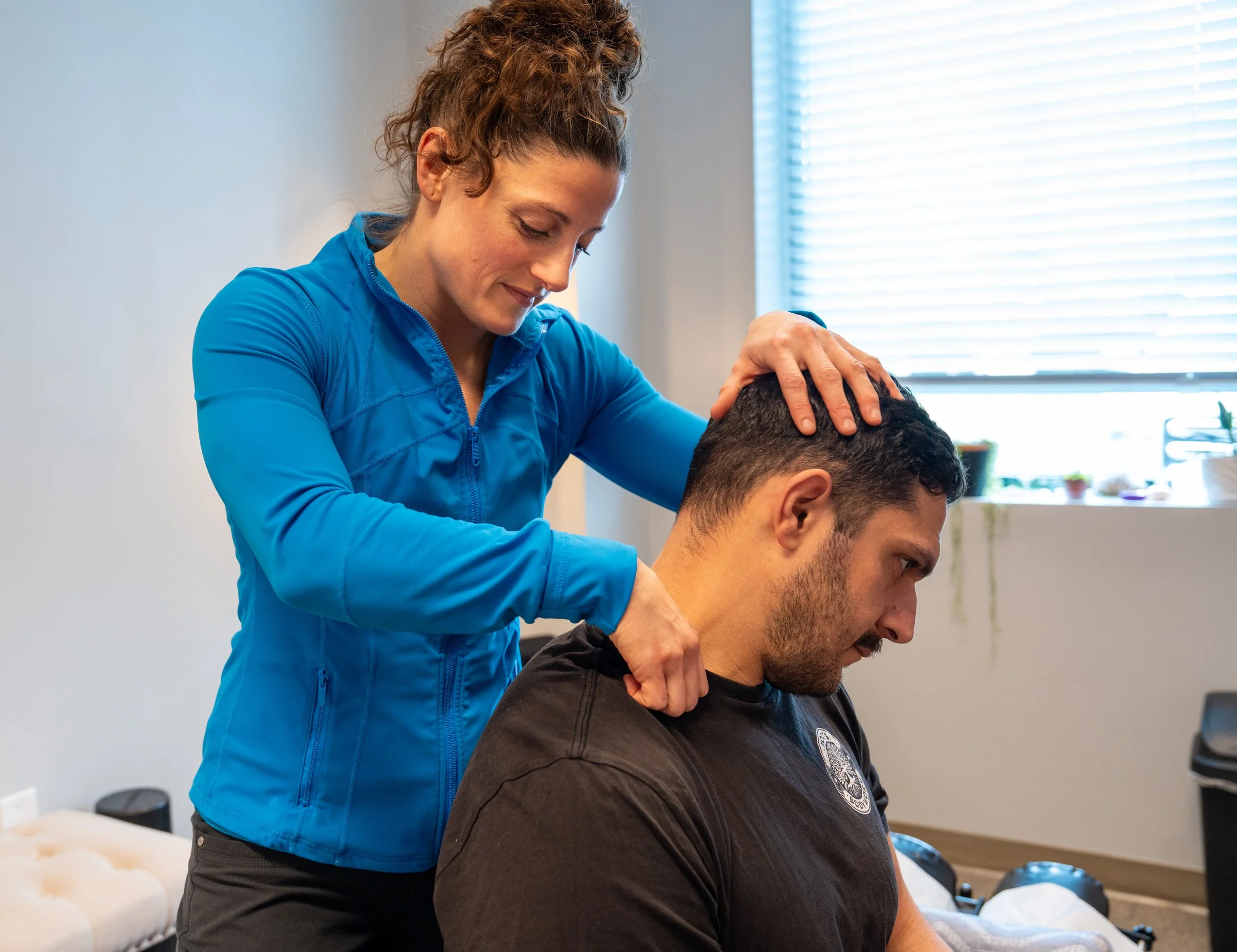 A woman in a blue athletic jacket provides chiropractic or massage therapy to a man who is sitting down with his head tilted forward, inside a room with a window and potted plants.