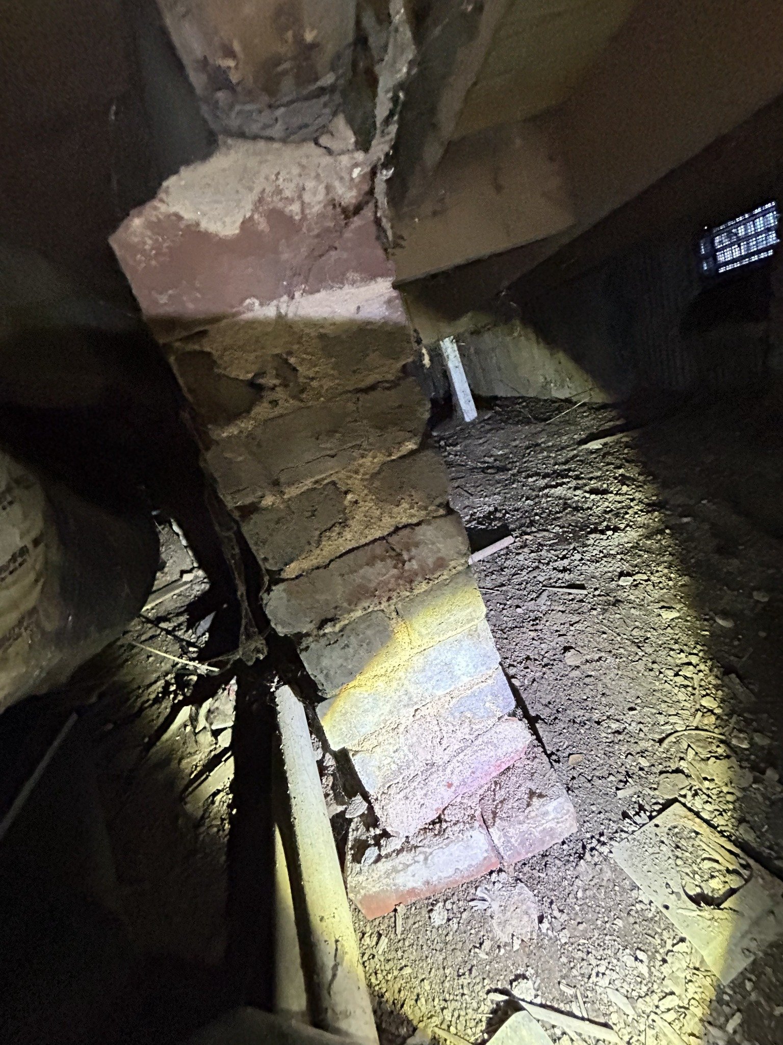 A close-up of a damaged brick wall with missing and broken bricks, with debris and dirt on the ground.
