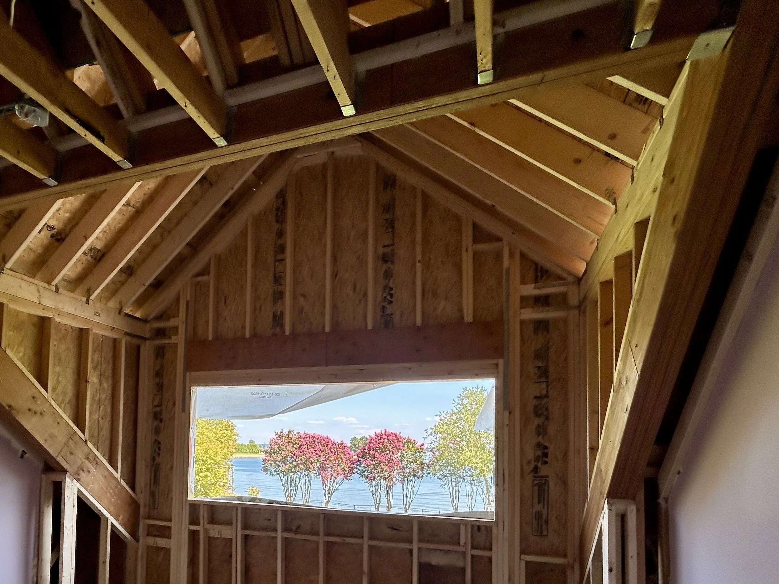 Interior of a building under construction with exposed wooden framing and an opening revealing a view of trees, water, and a clear sky outside.