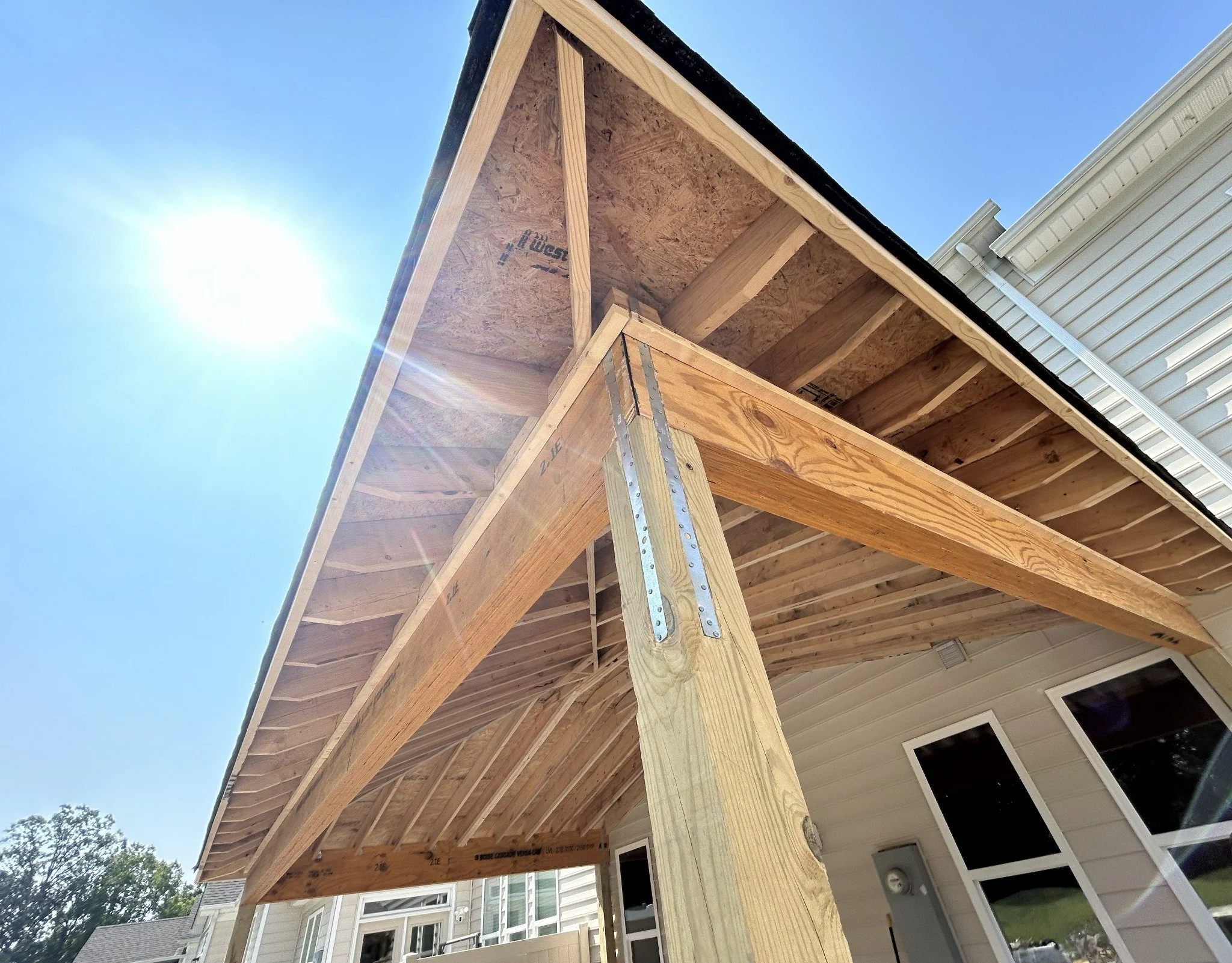 A building under construction with wooden framing and an overhanging roof, outdoors under a clear blue sky with sunlight, neighboring houses are visible in the background.