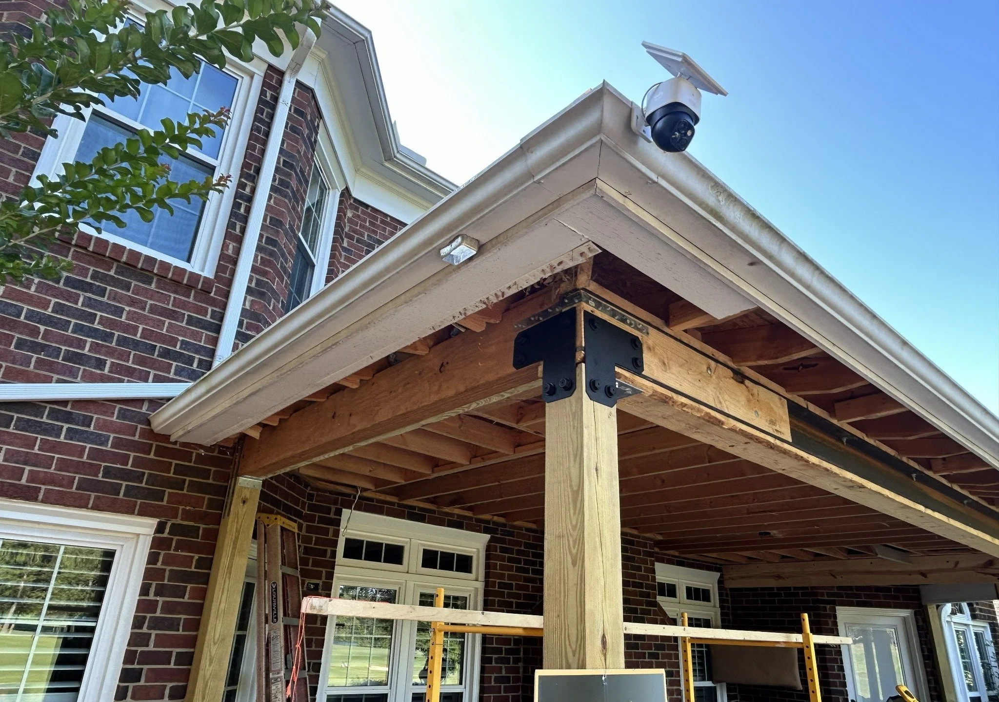 A house exterior under construction with brick walls and a partially built porch roof supported by wooden beams, a security camera installed on the house corner, and a clear blue sky.