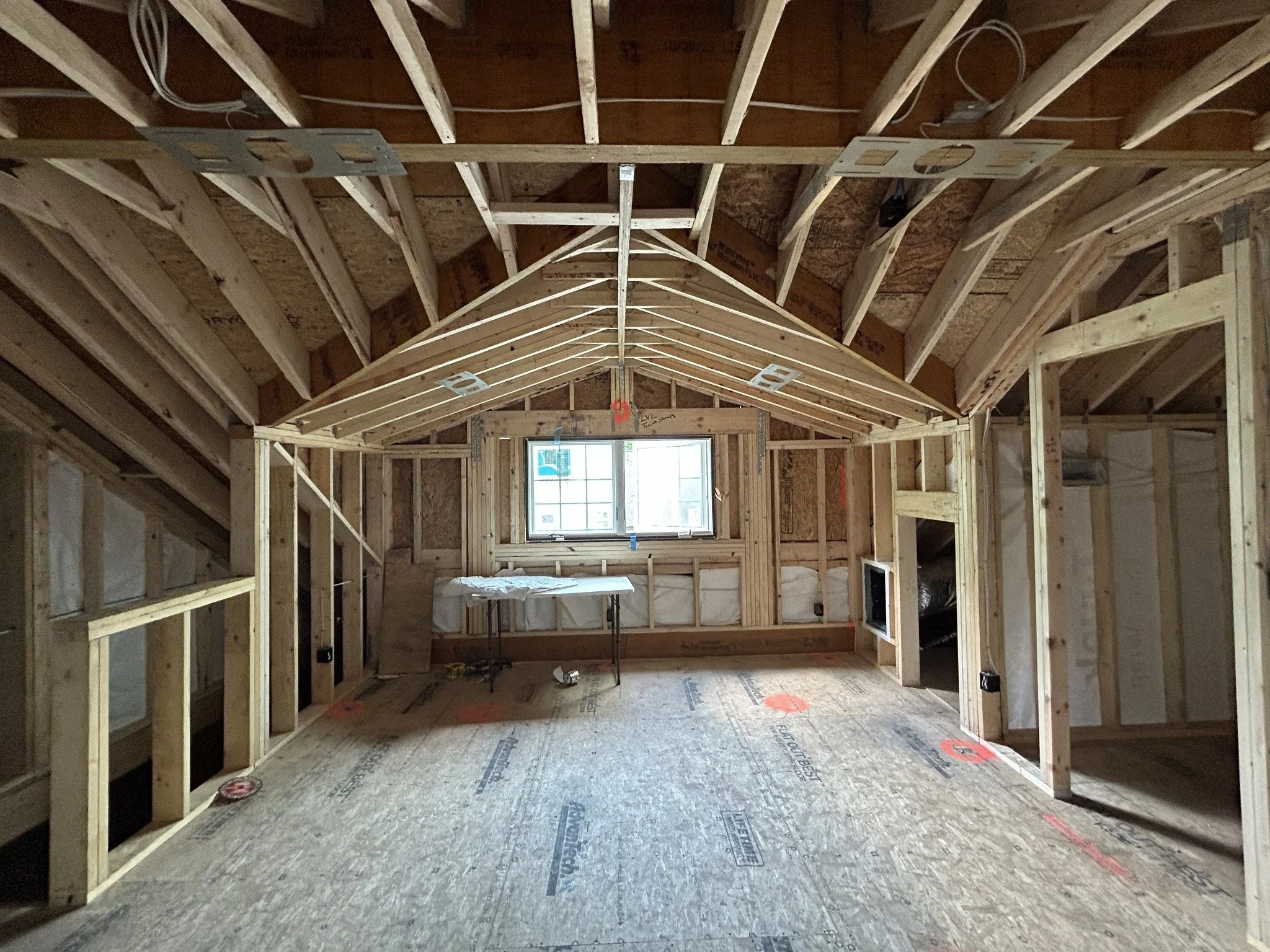 Interior view of a house under construction with exposed wooden framing, open wall spaces, and a small window at the end of the room.