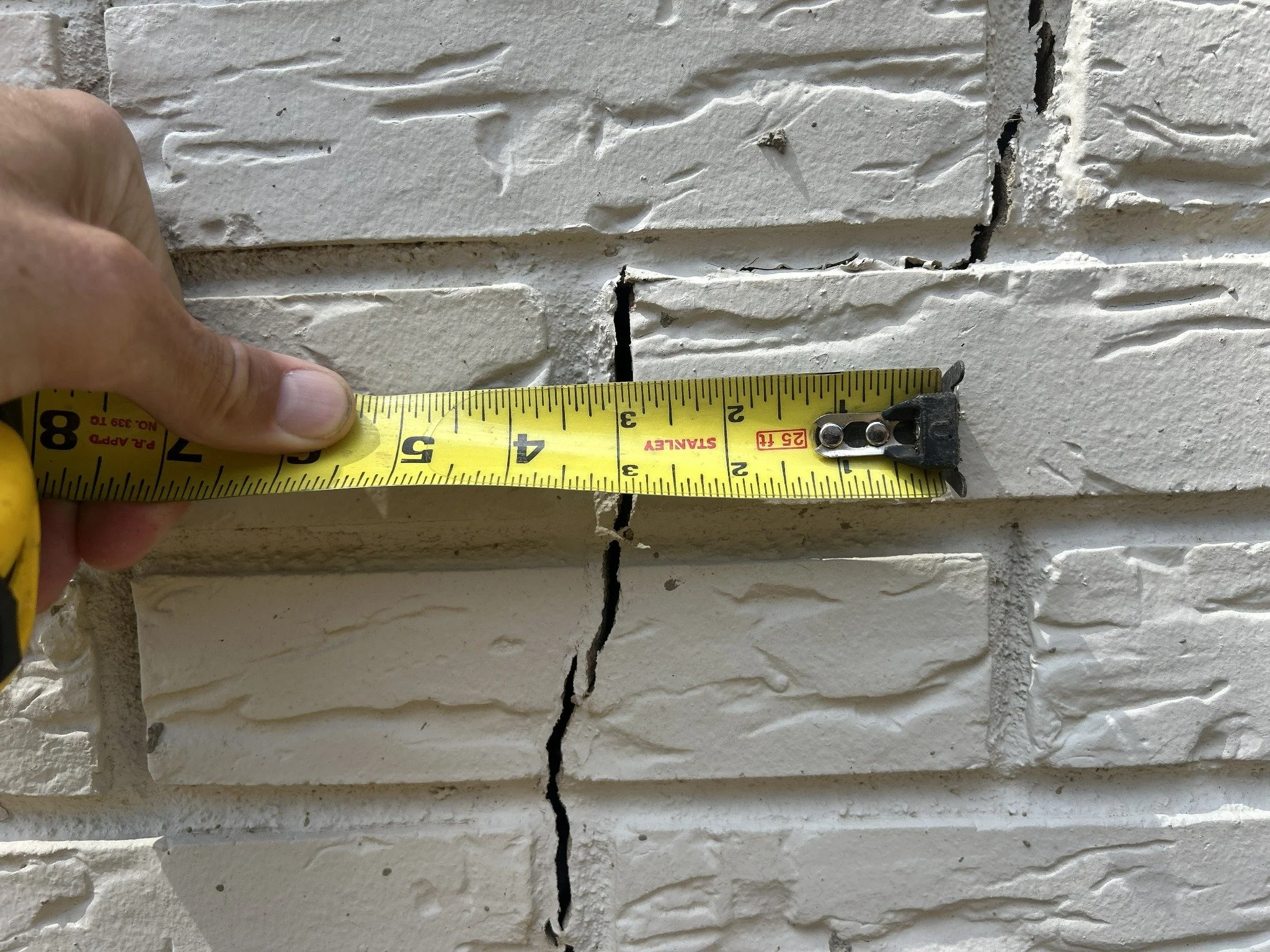 Close-up of a person's hand holding a yellow measuring tape against a white brick wall with a large crack in the mortar.