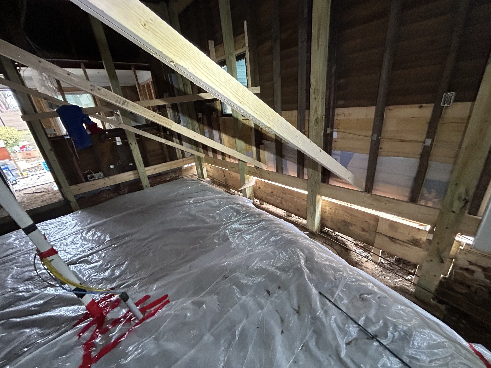 Interior of a house under construction with exposed wall studs and roof framing. There is a plastic sheet on the floor and some electrical wiring, with a window showing a neighborhood outside.