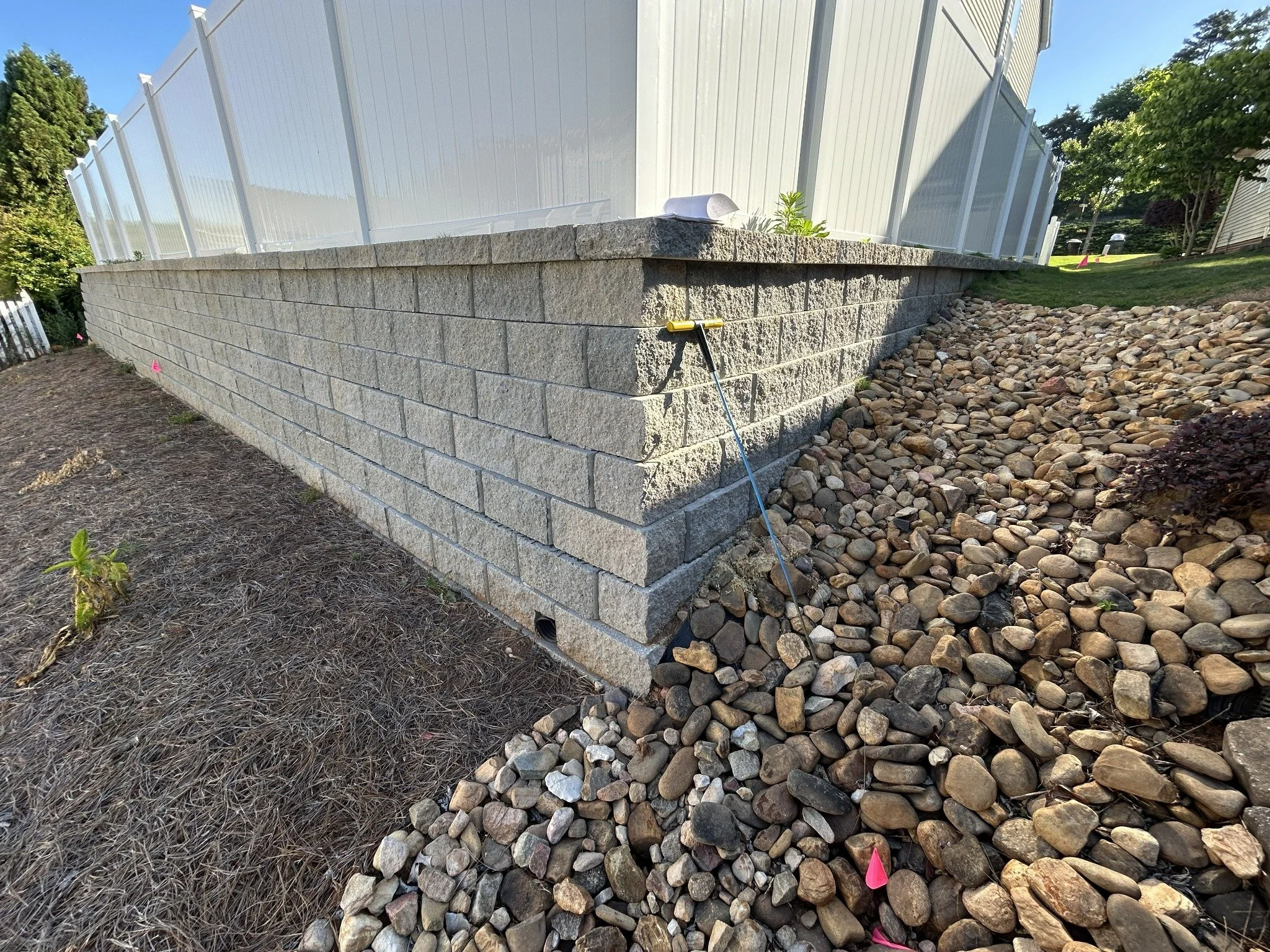 Photo of a stone retaining wall with gravel on one side and dirt with plants on the other, next to a white fence and a house, under a clear blue sky.