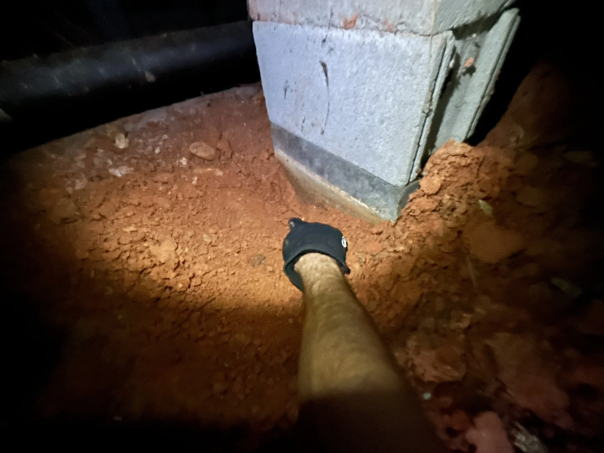 A person with a black watch on their wrist is taking a photo of their leg inside a construction or renovation area. The ground is dirt, and there is a concrete foundation or wall with some exposed bricks.