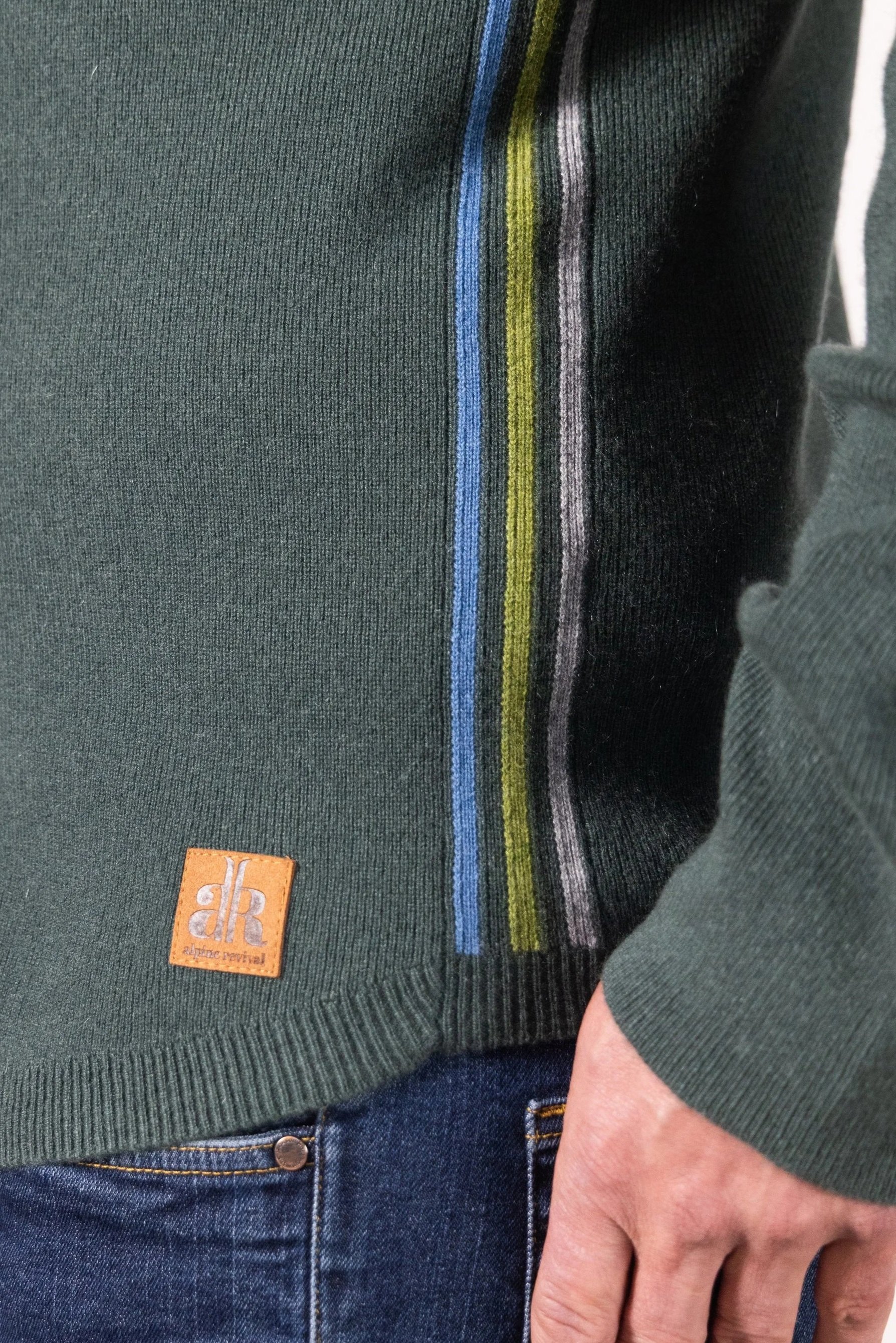 A man wearing a dark green button-front sweater with 2 chest pockets, a shirt-style collar, a yoke similar to a western shirt and multicolored stripes down the sides, his hands are in his pants pockets and he is smiling.