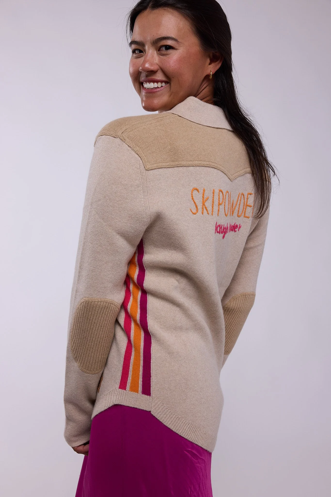 A woman smiling wearing a vibrant pink hoodie sweater with striped details on the sides, drawstrings at the hood and a cozy cowl neck with jeans.