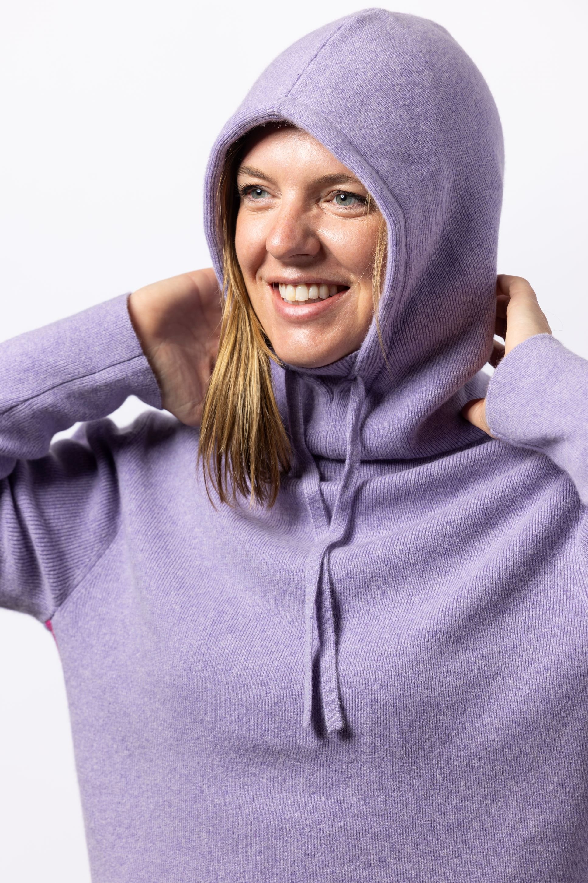 Woman in a studio wearing a lavender cashmere hoodie with retro stripes on sides with hoodie pulled up over her head