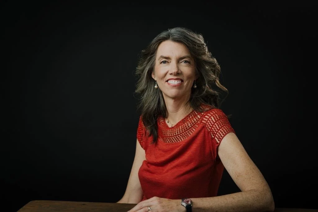 A woman with long, wavy brown hair wearing a red embroidered top, sitting at a wooden table against a black background, smiling at the camera.