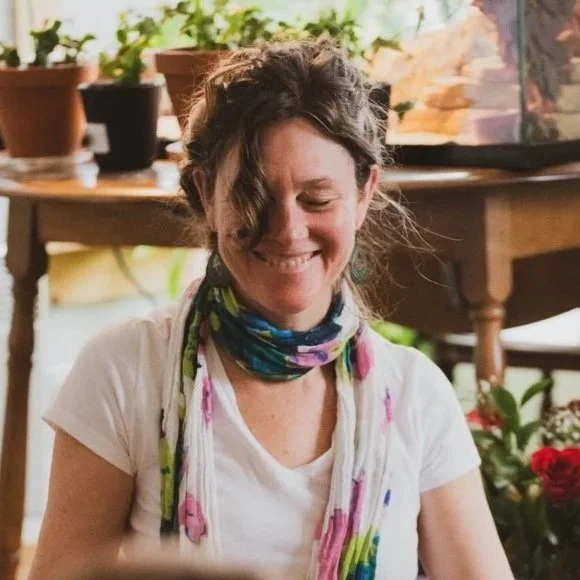 A woman with curly hair smiling, wearing a white shirt and a colorful scarf, sitting indoors with potted plants and wooden furniture in the background.