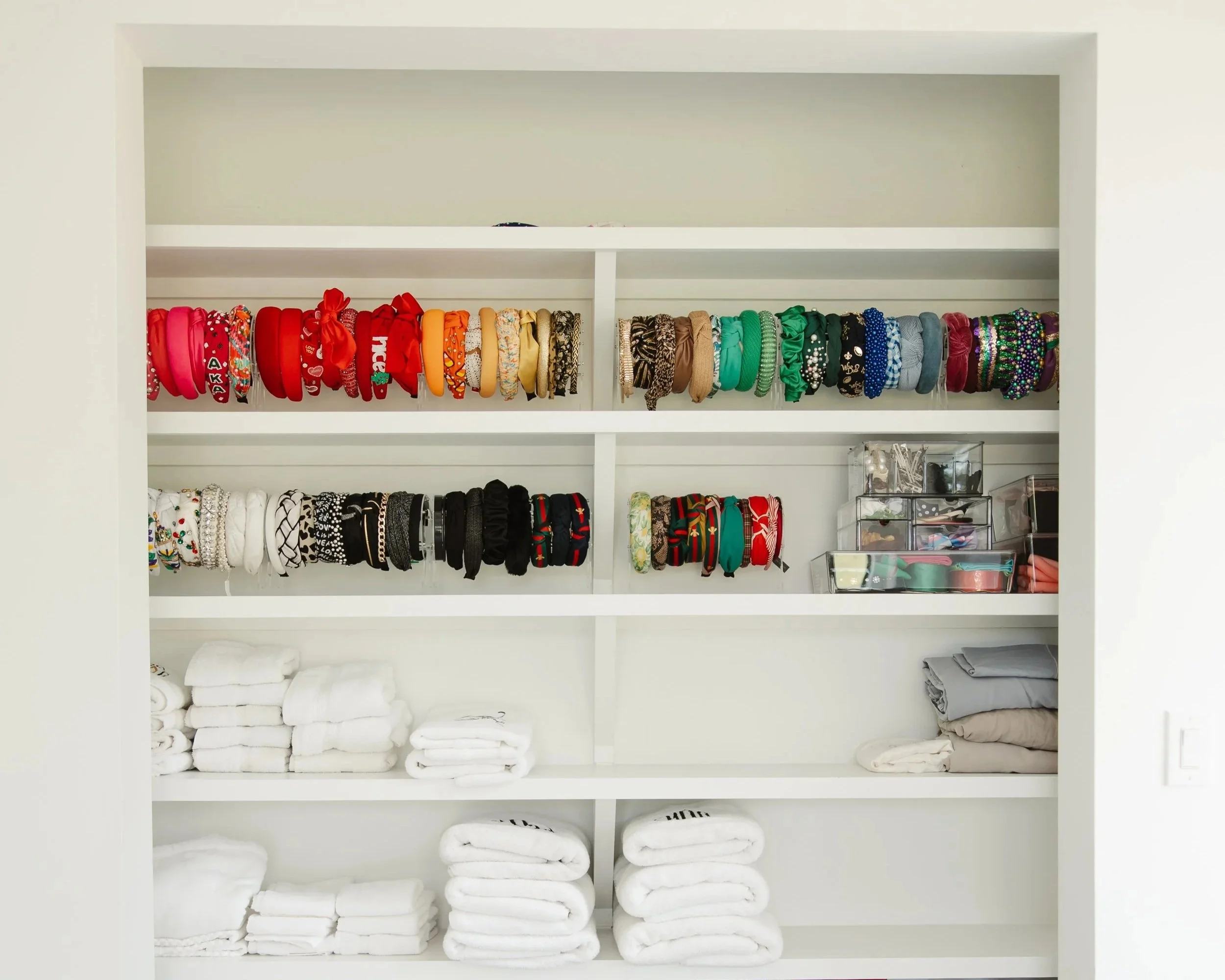 a bathroom shelf that shows white neatly folded towels and organizers that show headbands being displayed in a rainbow color fashion