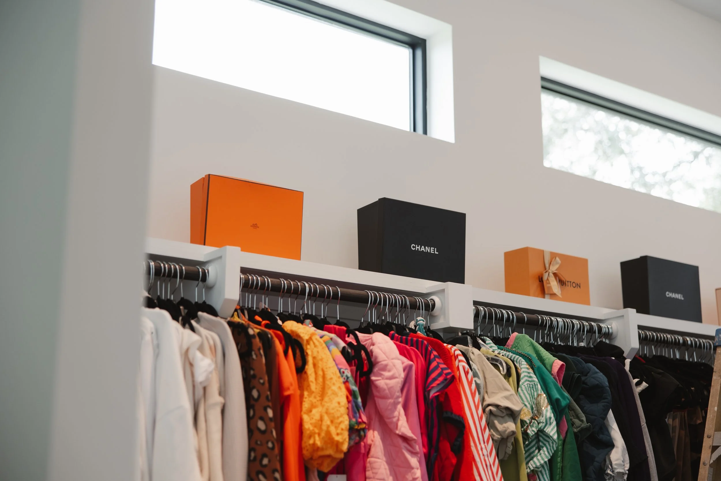 a closet rack that has colored-coded shirts displayed and on a shelf luxury brand boxes like chanel, hermes, and louis vuitton
