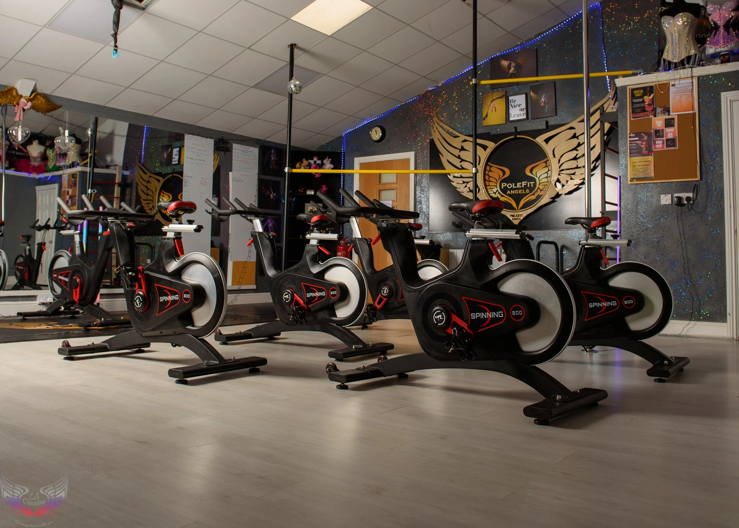 Gym classroom with multiple stationary exercise bikes arranged in a row, a colorful wall with artwork, a mirror reflecting part of the room, and fitness-related decoration.