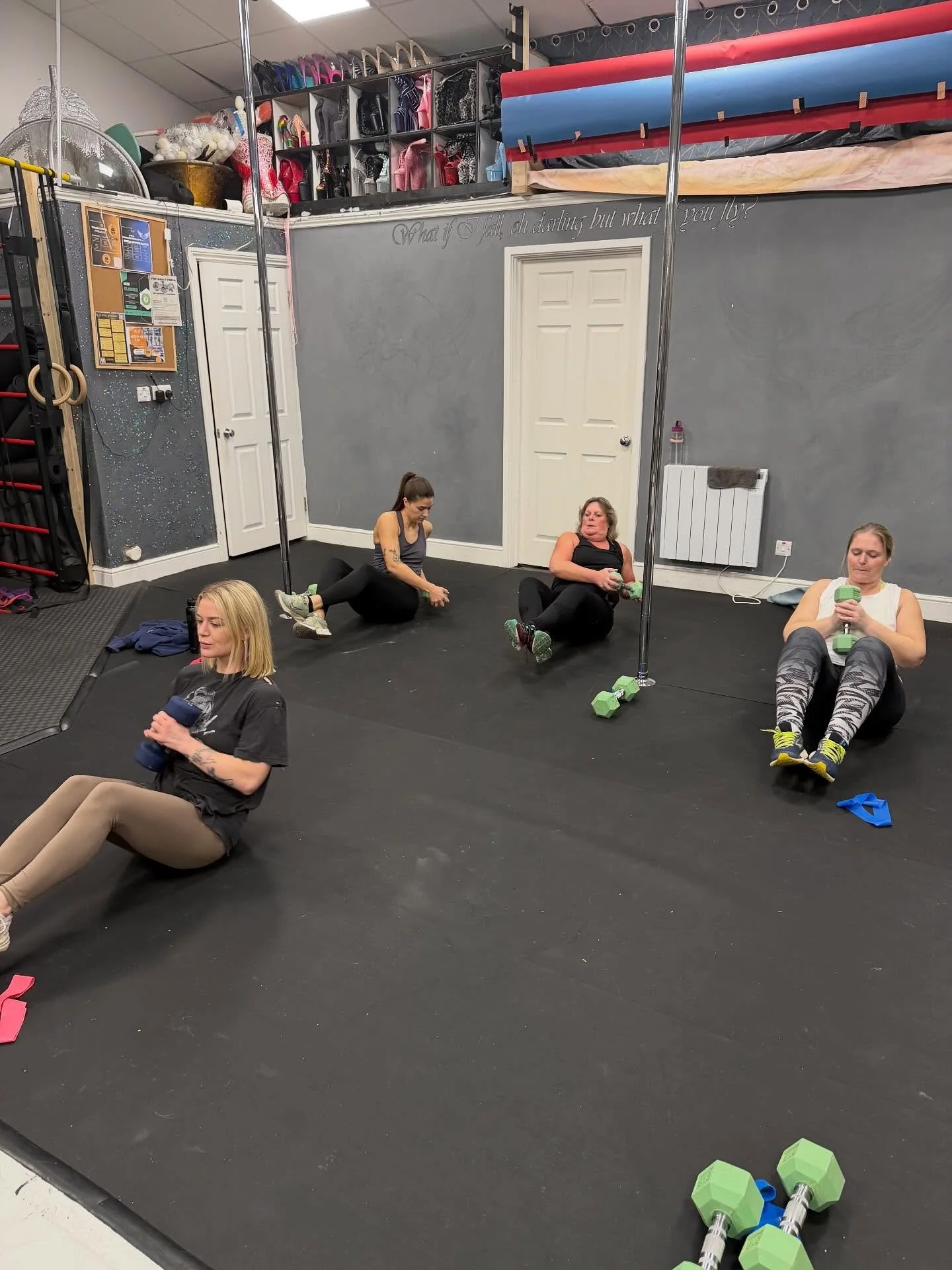 Four women are sitting and lying on the floor of a gym, leaning against a mirrored wall, during a workout session. They have dumbbells and are using their phones or resting. The background shows gym equipment, storage shelves, and a racetrack on the ceiling.