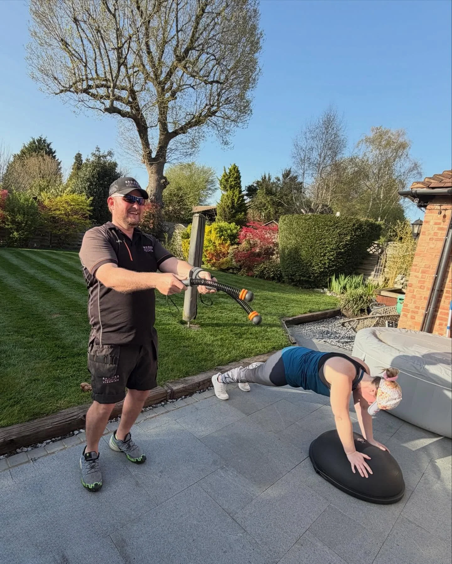 A man is chipping a woman who is doing push-ups on a balance board in a backyard with green grass, trees, and bushes under a clear blue sky.