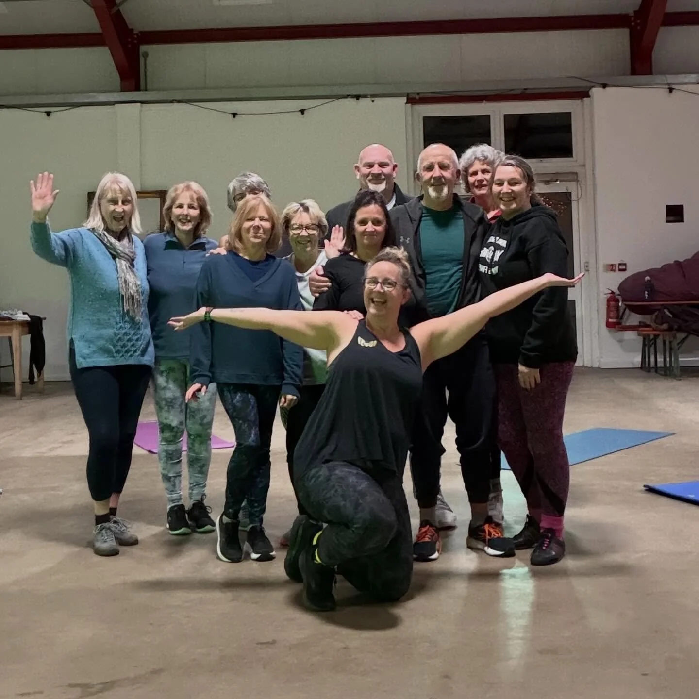 A group of ten people posing indoors in a spacious hall, with one woman kneeling in the front with arms outstretched and smiling, and the others standing behind her, some waving and smiling.