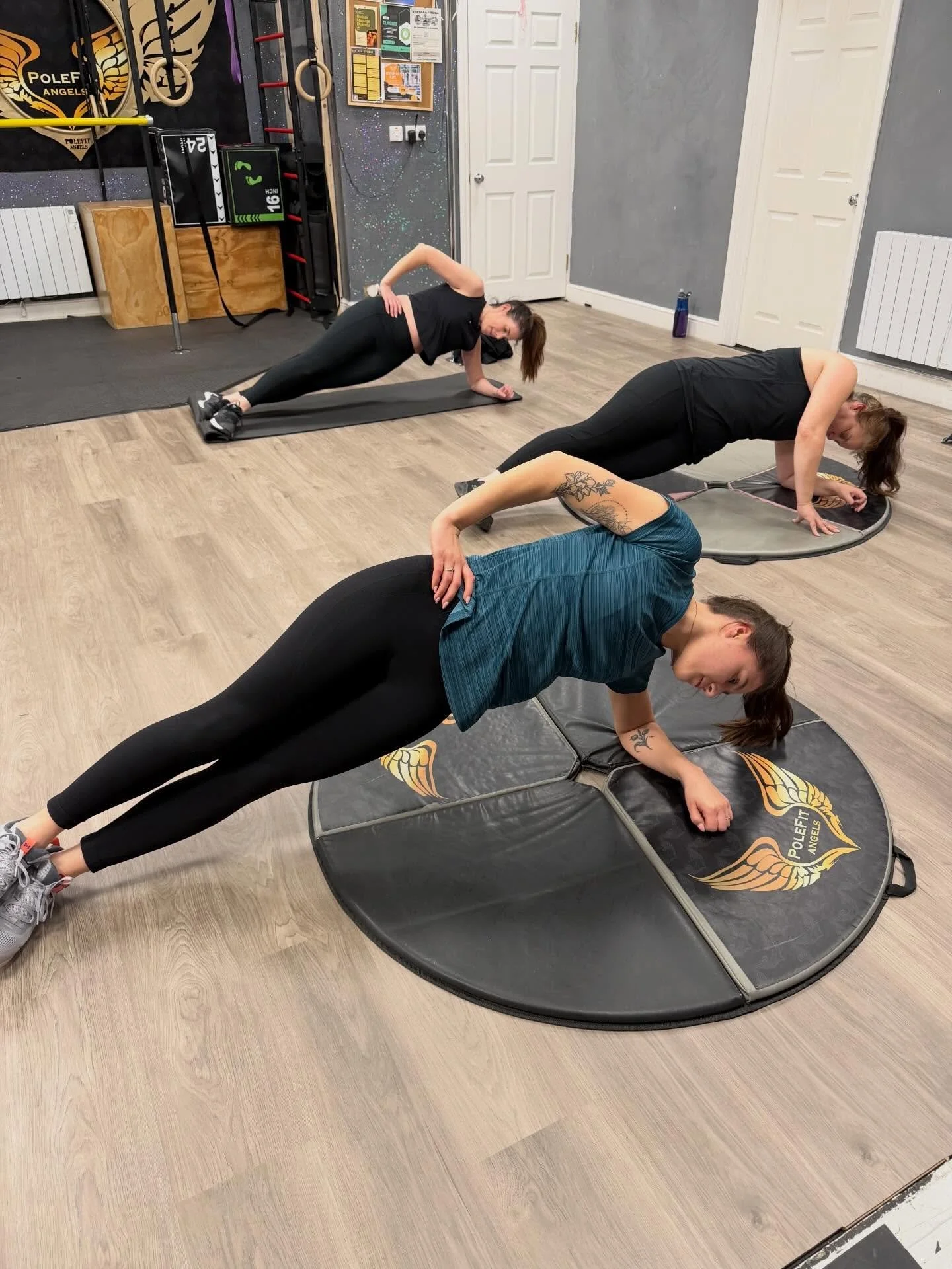 Three women exercising on black workout mats in a gym, performing side planks with their bodies lifted and supported on one arm.