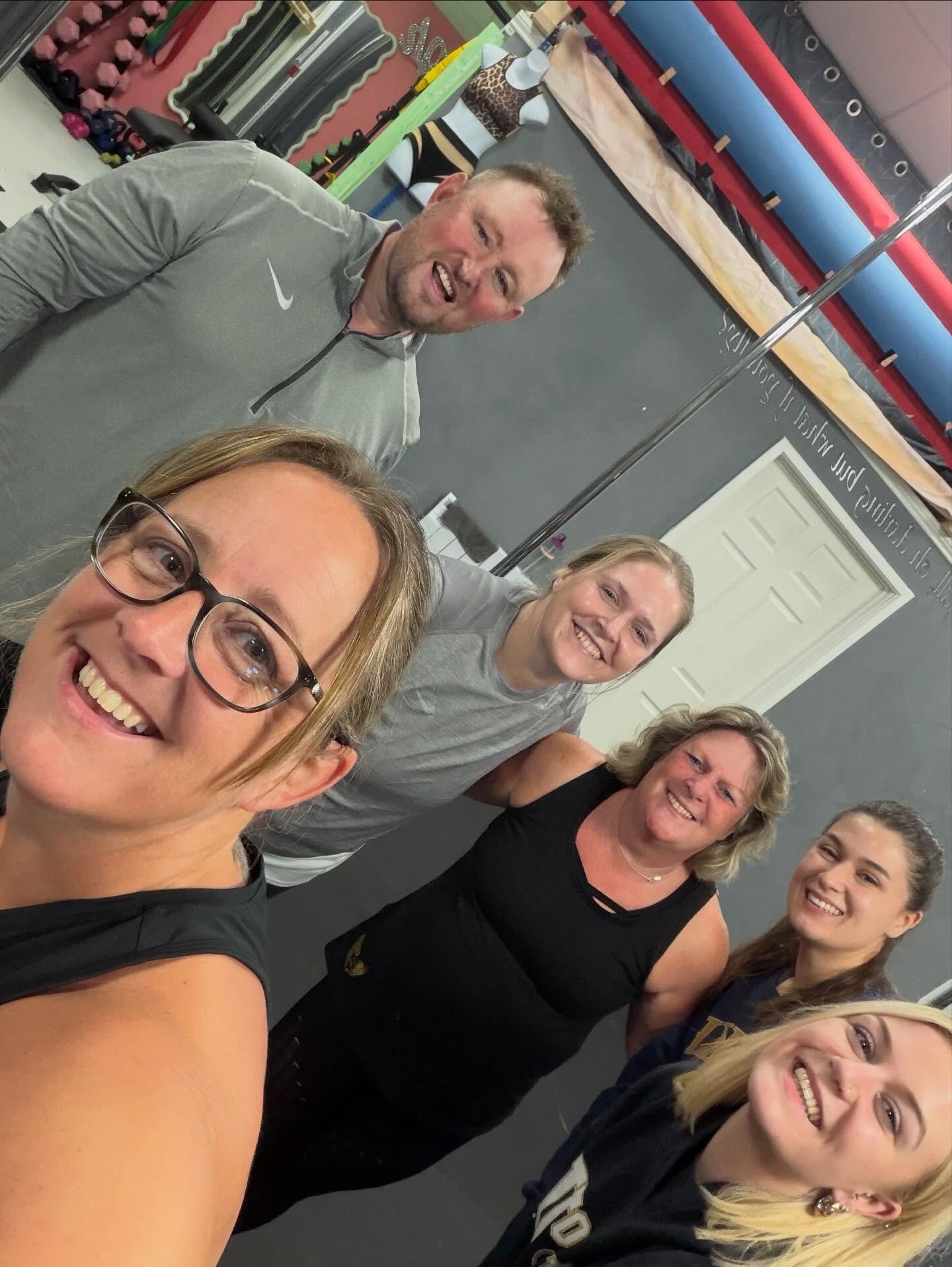 Group of six smiling people taking a selfie in a gym, with workout equipment and mats in the background.