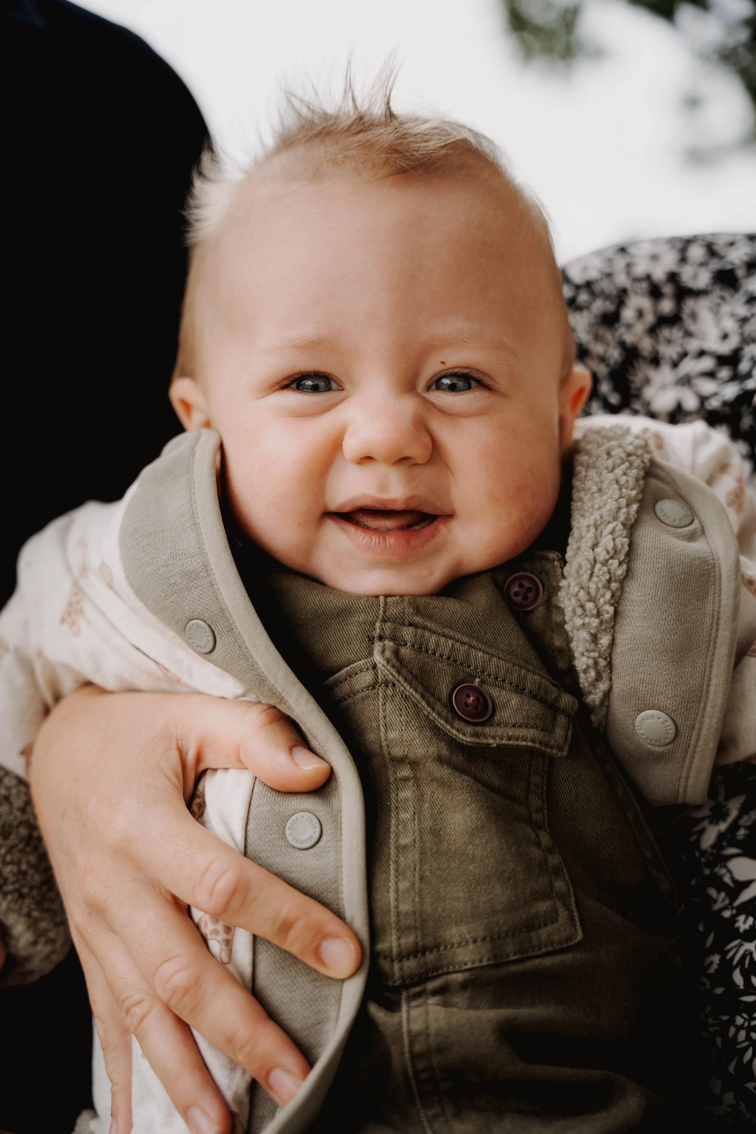 Close-up of a smiling baby with light brown hair and blue eyes, wearing a beige and green vest, held by an adult.