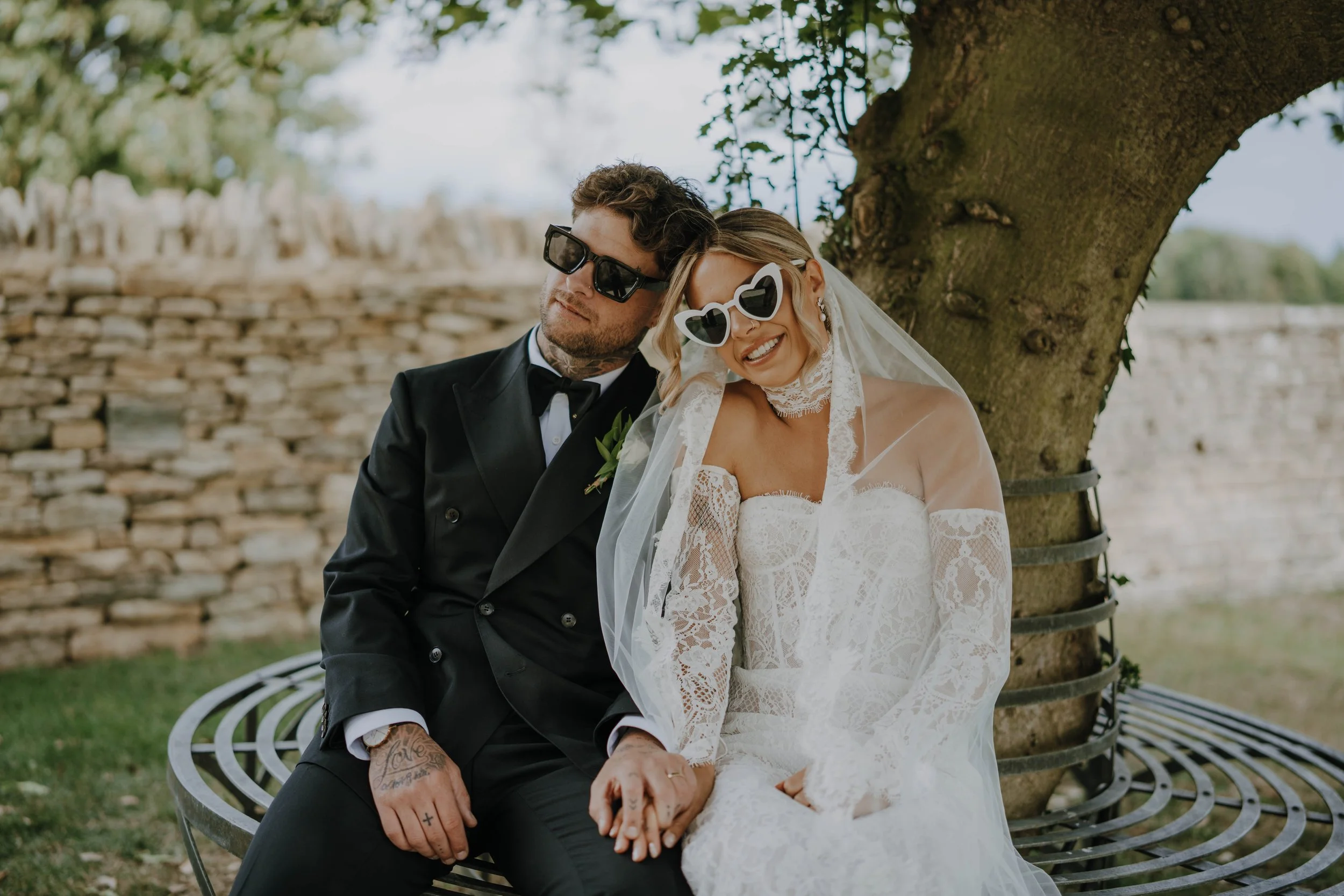 A bride and groom sitting on a bench under a tree, holding hands, both wearing sunglasses, with a stone wall in the background.