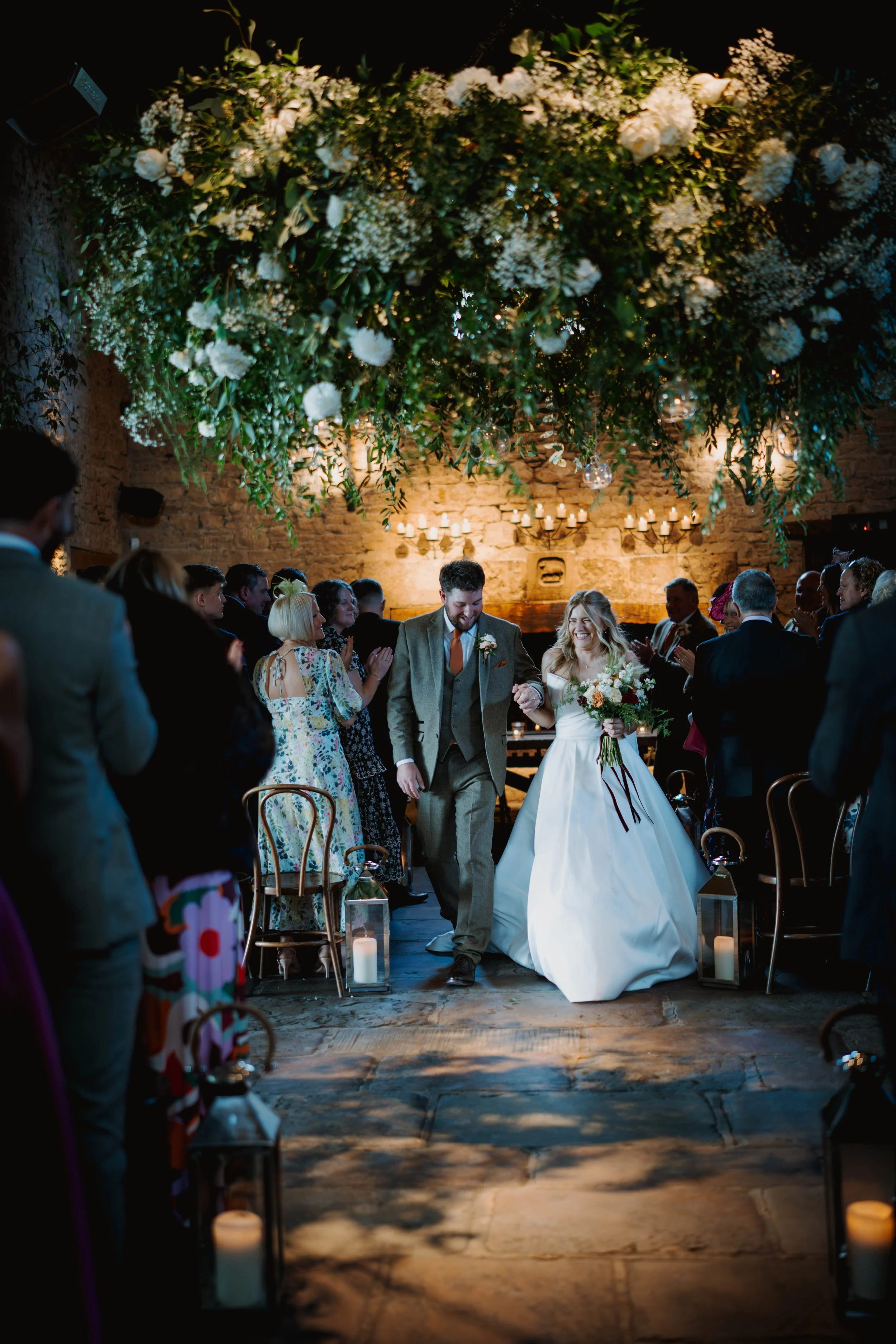 A bride and groom are walking hand in hand at their wedding reception, smiling and surrounded by guests. The ceiling is decorated with lush greenery and white flowers, creating an elegant ambiance. Candles in lanterns line the floor, and warm lighting illuminates the rustic brick wall in the background.