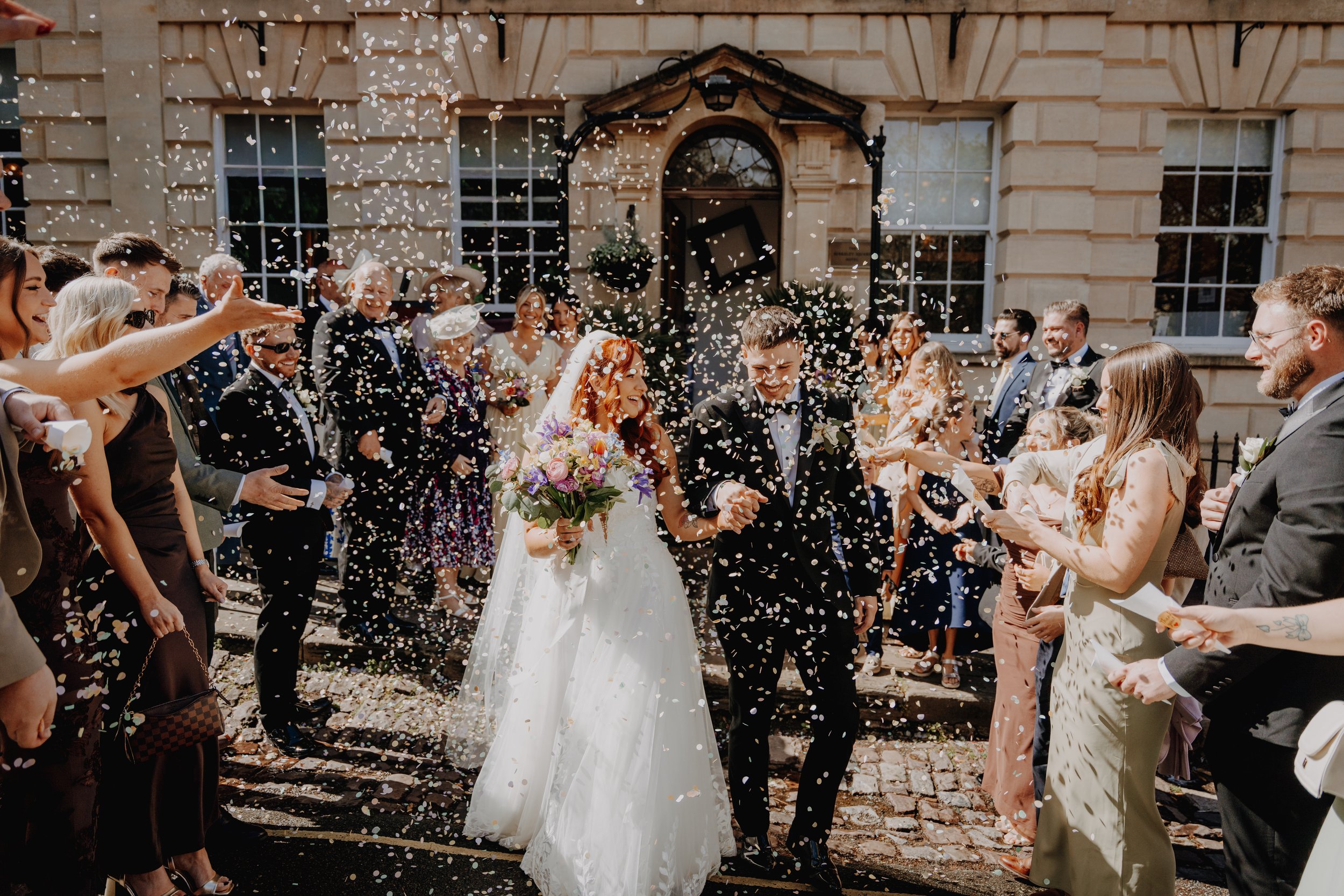 A wedding celebration with a bride and groom walking through a crowd outside a building, surrounded by guests throwing confetti.