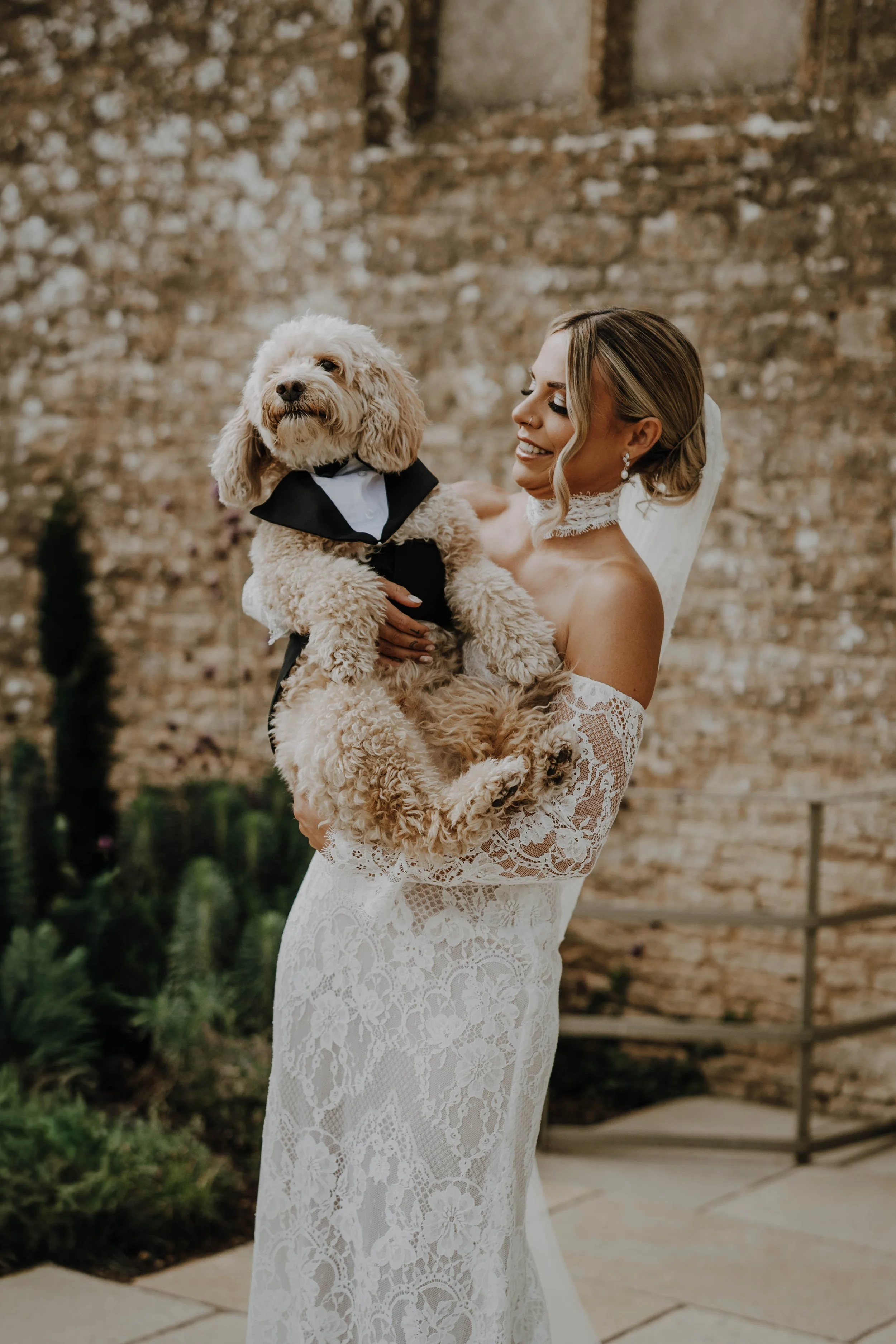 A bride in a white lace wedding dress holding a dog dressed in a tuxedo, standing outdoors against a brick wall.