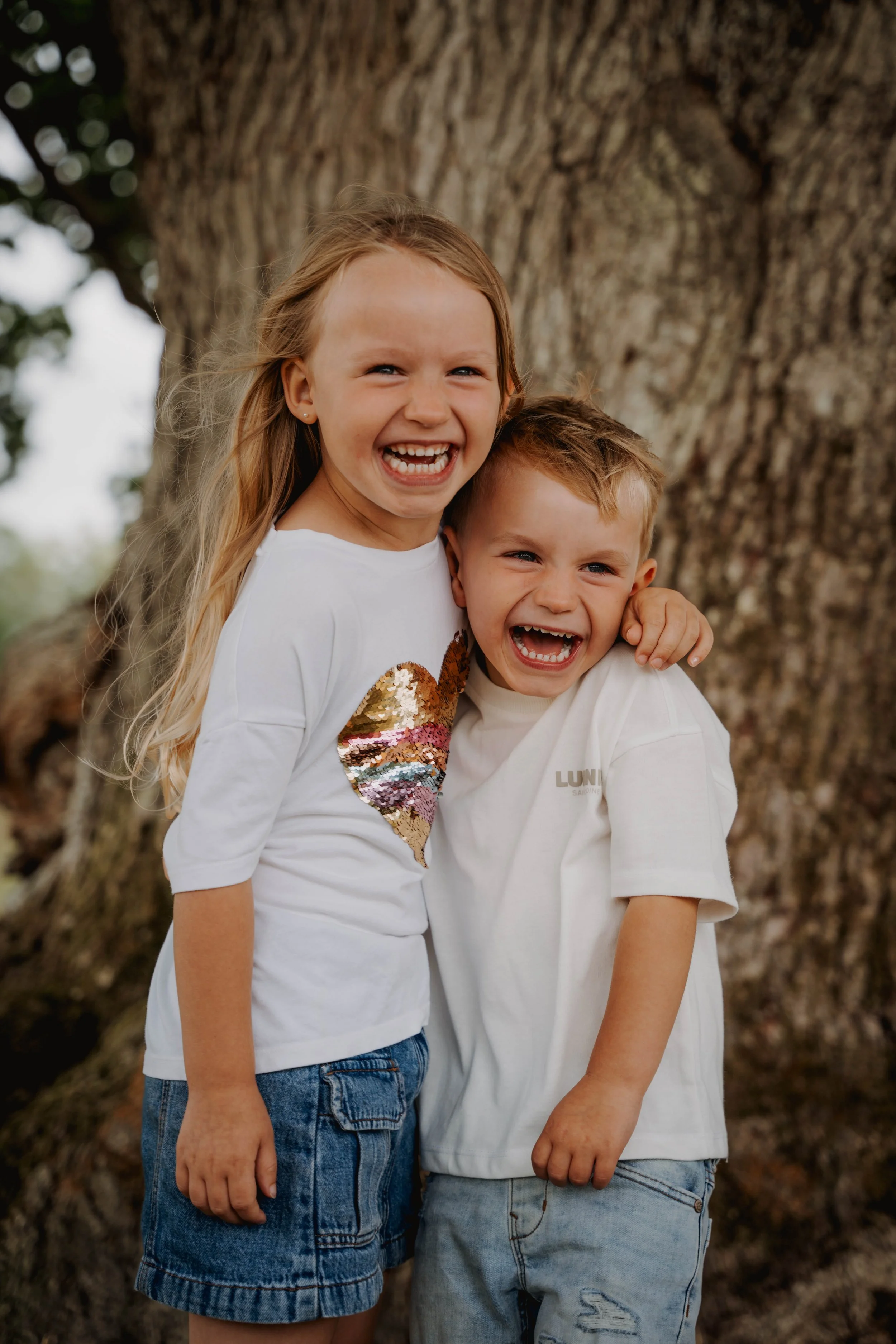 Two children, a girl and a boy, smiling and hugging each other in front of a large tree trunk.