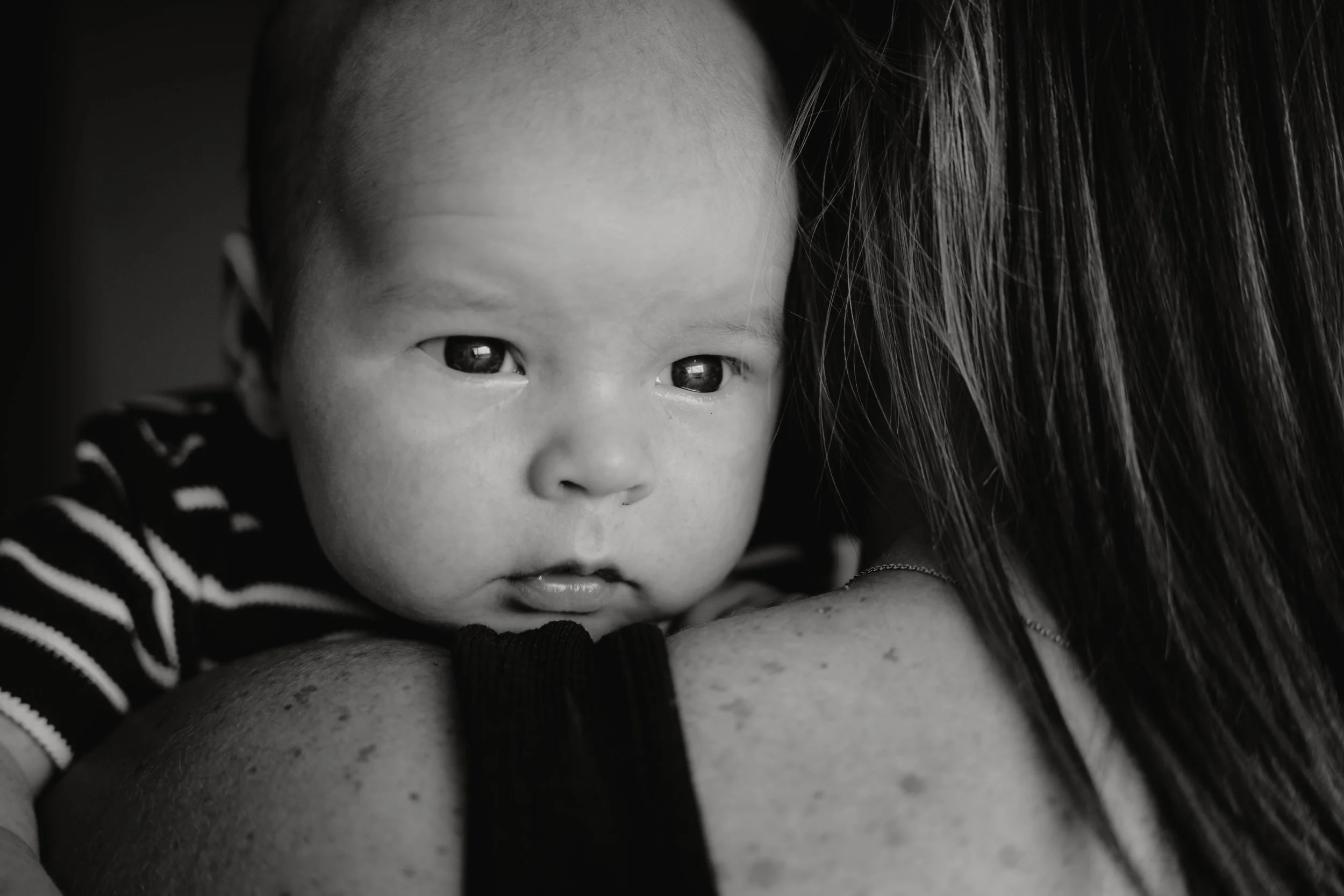 Black and white photo of a baby with Asian features resting on a person's shoulder, looking directly at the camera.