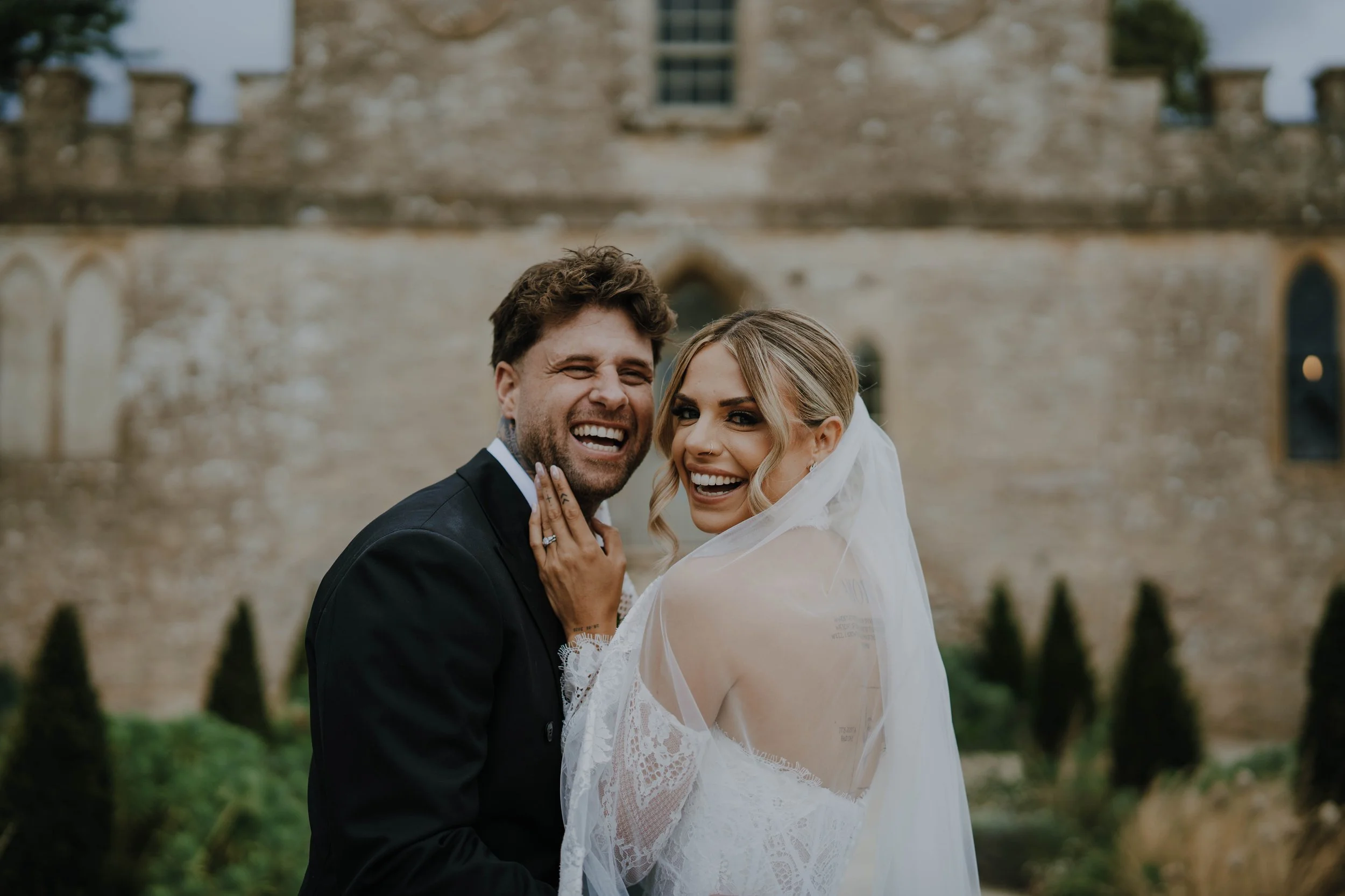 A bride and groom smiling in wedding attire outdoors, with a castle-like stone building in the background.