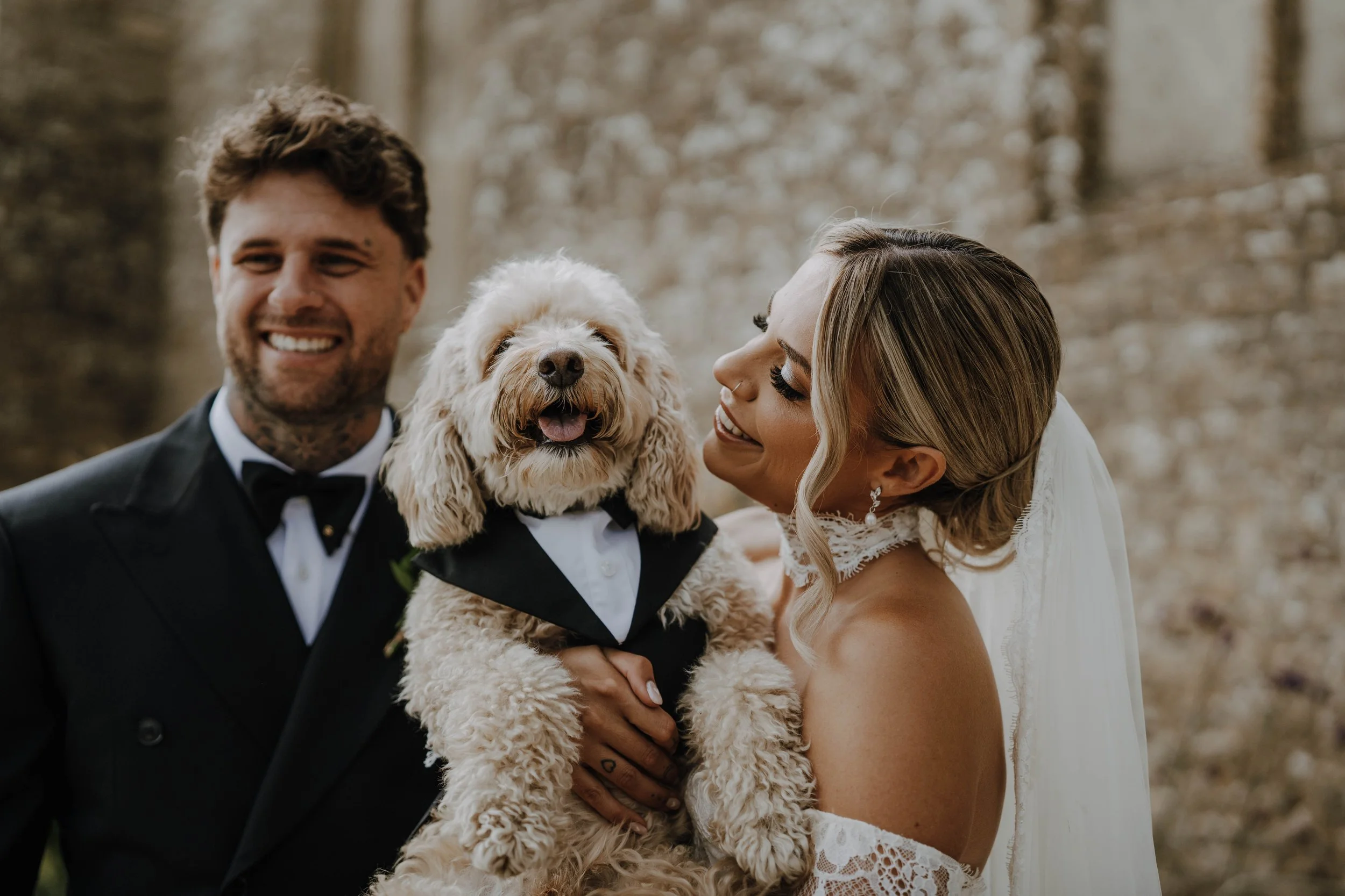 A bride, groom, and a woman holding a fluffy dog dressed in a tuxedo with a bow tie, in an outdoor setting with a blurred natural background.