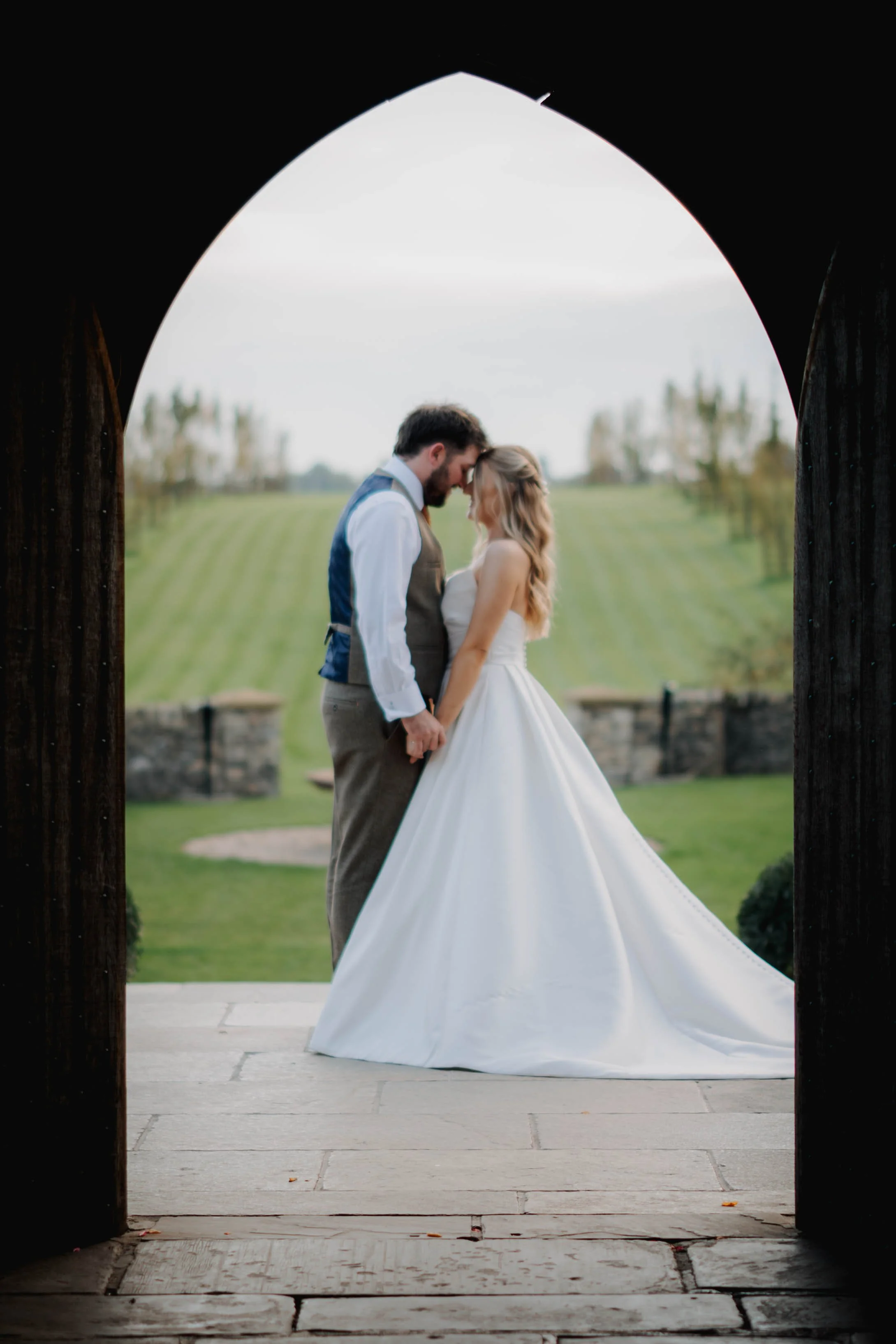 A bride and groom standing close, touching foreheads and holding hands under a wooden archway outdoors. The bride is in a white wedding gown, and the groom is in a vest and dress shirt. The background features a grassy landscape with scattered trees.