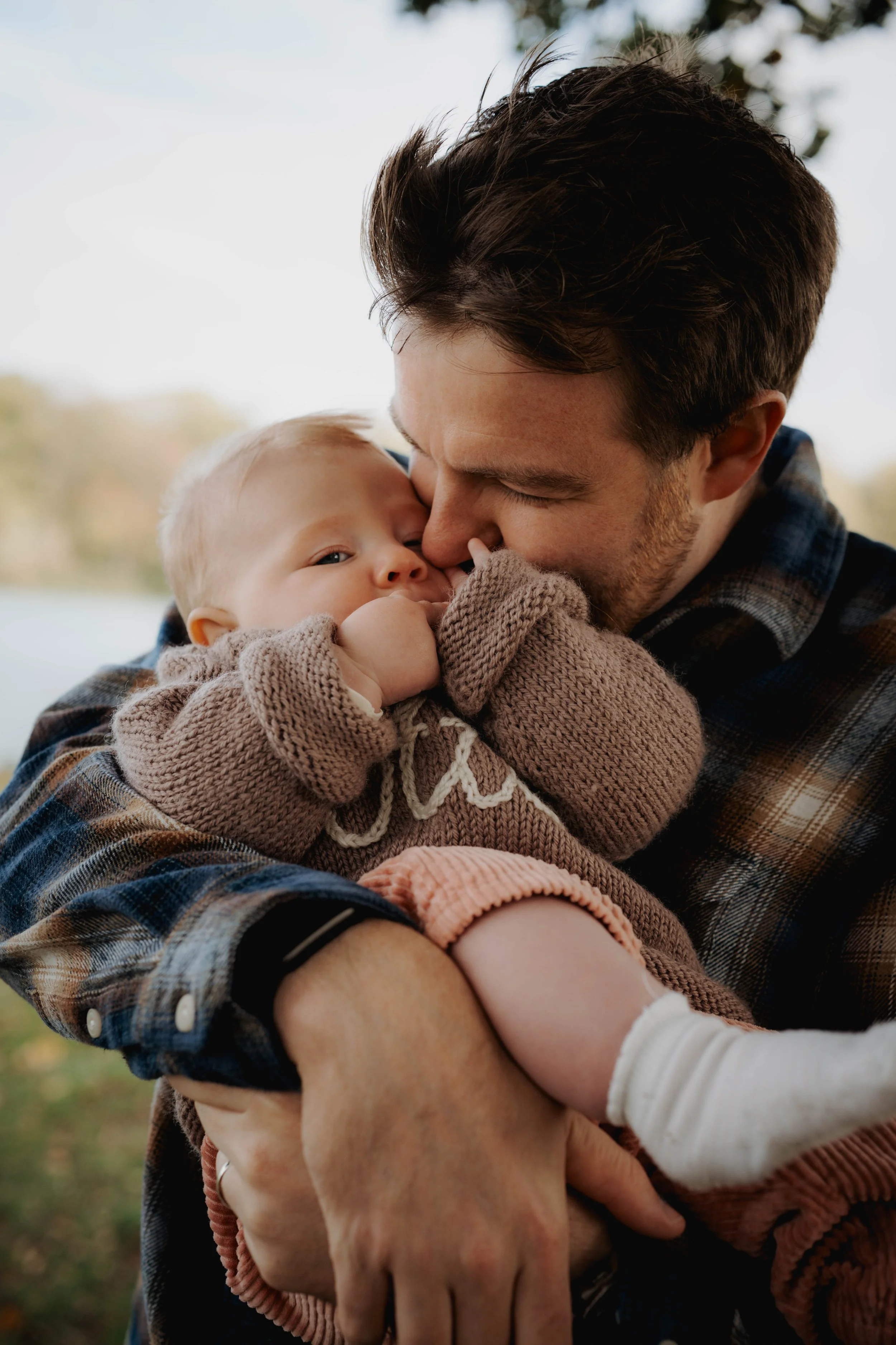 A man holding a baby close to his face, both with their eyes closed, sharing a tender moment outdoors on a bright day.
