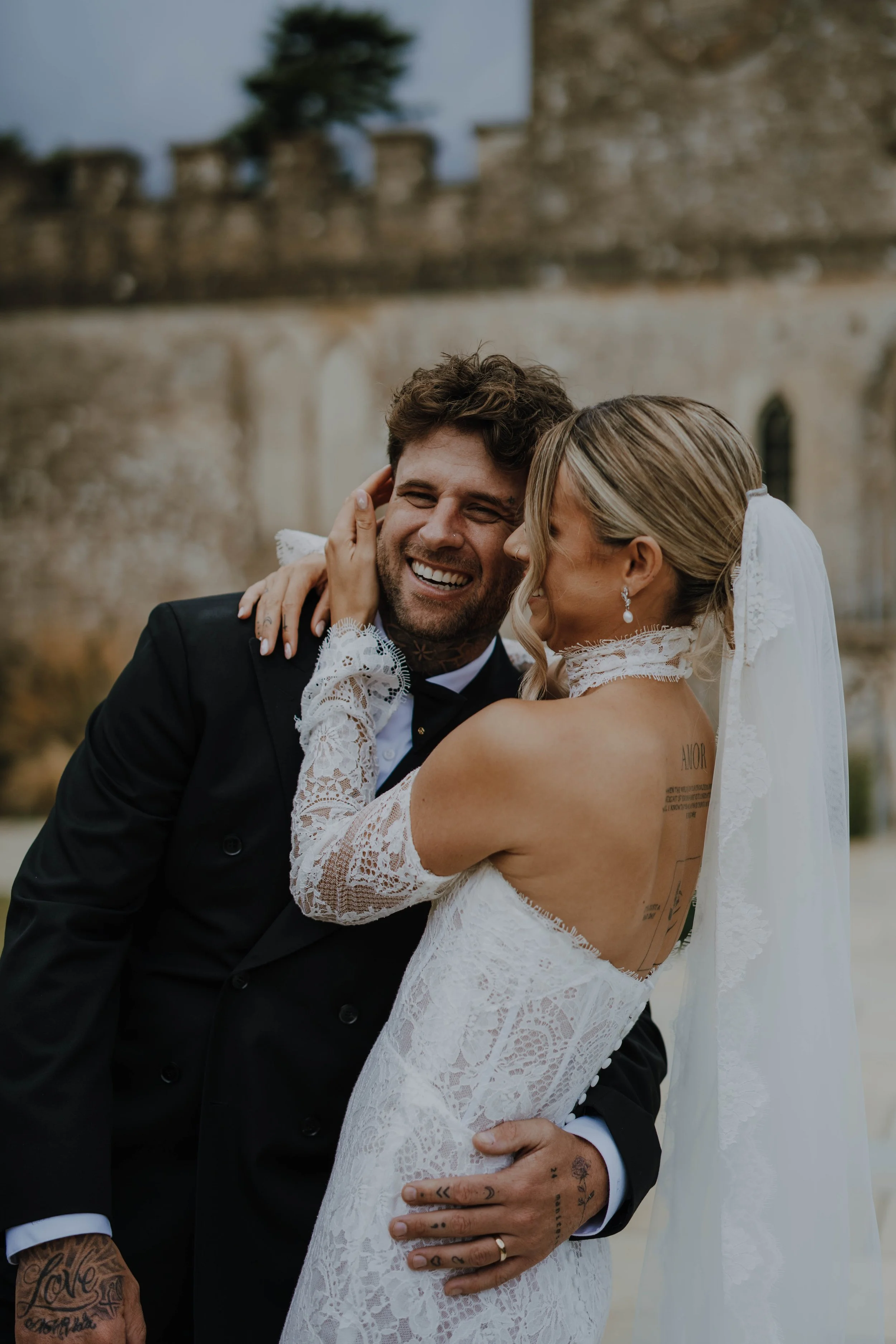 A smiling couple dressed in wedding attire, with the bride in a lace gown and veil, embracing outdoors in front of a stone building with gothic windows and a stone wall, sharing a joyful moment.