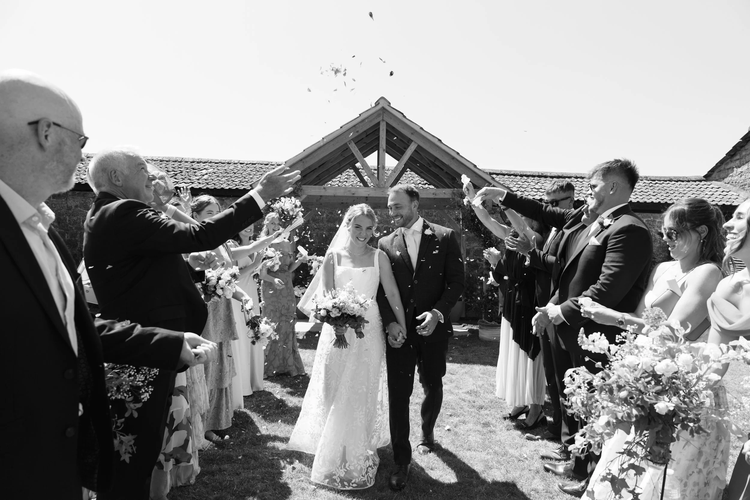 A black-and-white photo of a wedding celebration outdoors. The bride and groom hold hands at the center, smiling. Wedding guests form a corridor on both sides, throwing flower petals and cheering. A rustic building with a tiled roof and wooden beams 