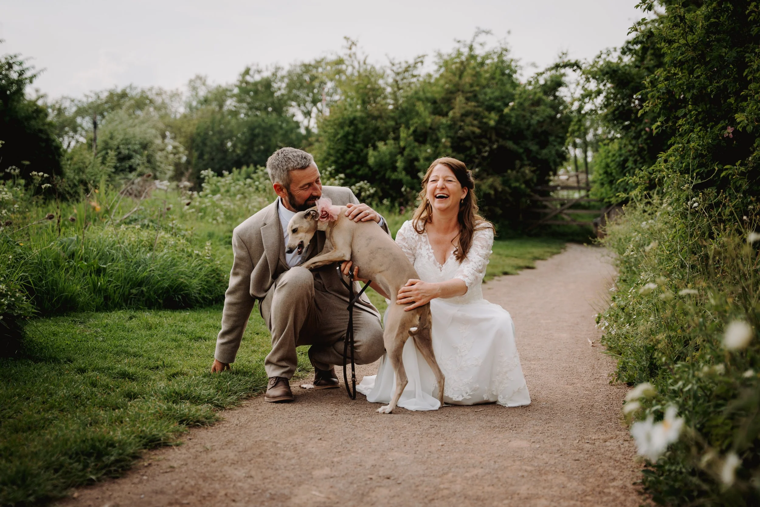 A man and woman in wedding attire spending joyful moment with a dog outdoors on a garden path surrounded by greenery.