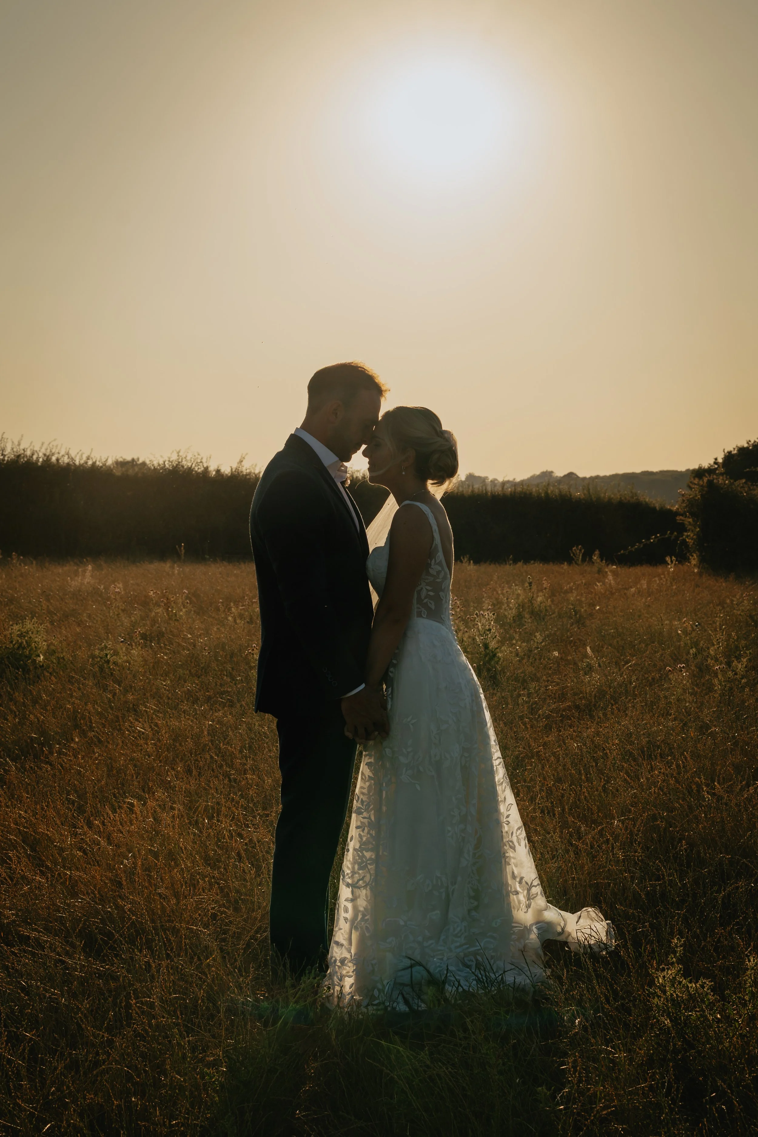 A silhouette of a bride and groom holding hands and leaning their foreheads together in a field during sunset.
