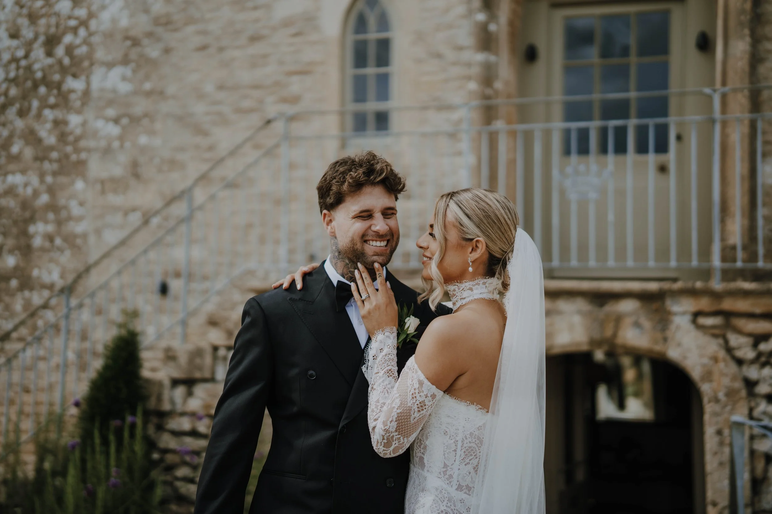 A bride and groom smiling and touching faces outdoors in front of a brick building, with the bride holding the groom's face.